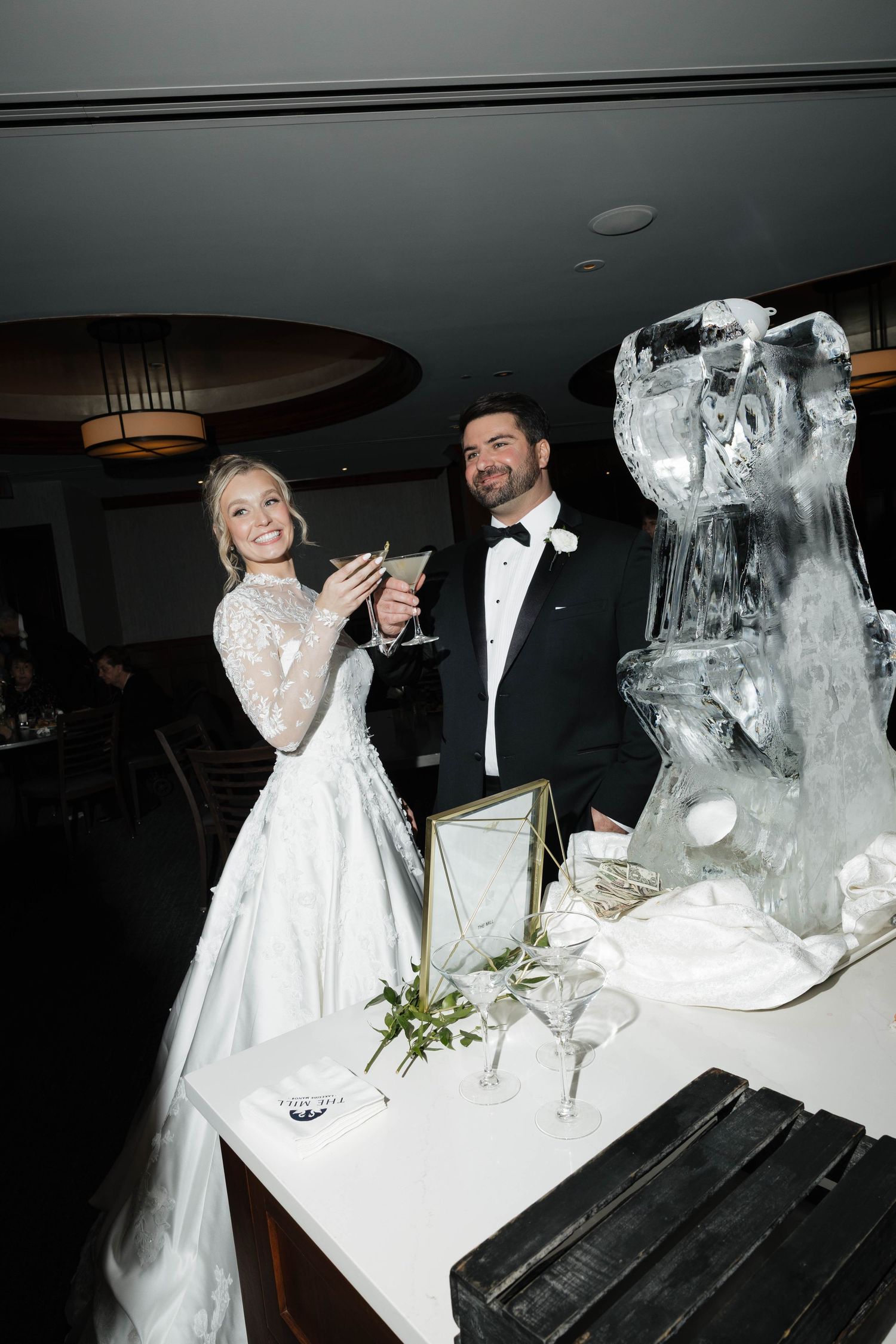 A bride and groom laugh while raising champagne glasses next to a decorative ice sculpture.
