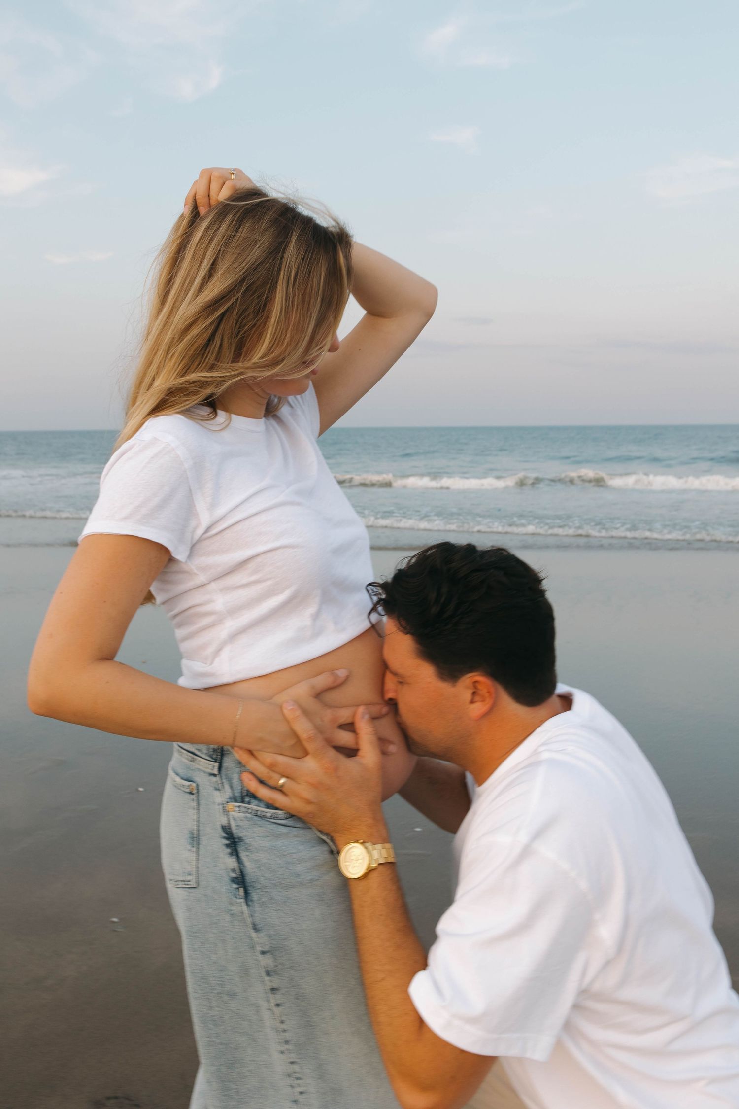 A couple shares an intimate moment on a beach at sunset with mountains in the background.