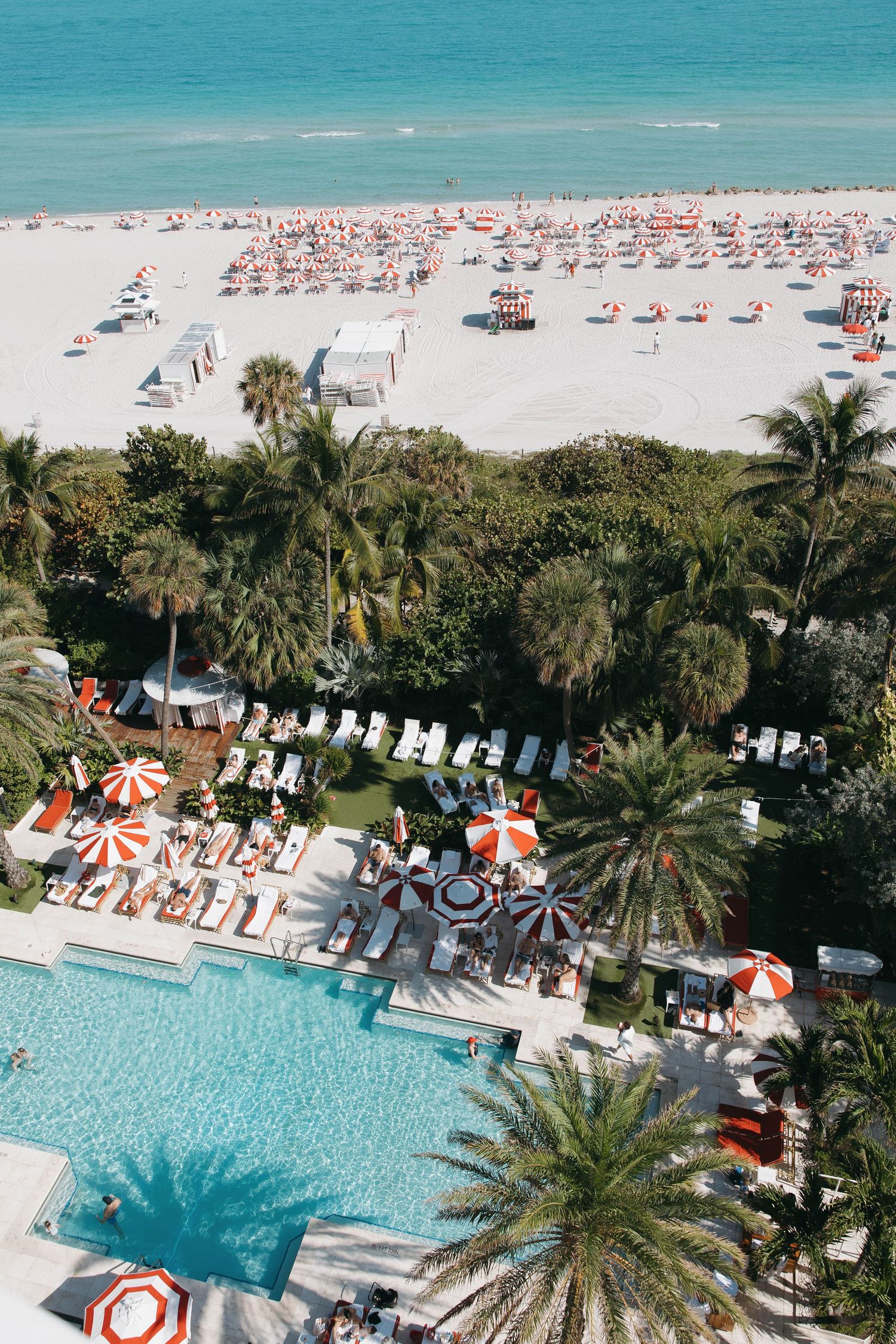 Aerial view of Miami Beach with turquoise ocean, white sand beach, palm trees and resort pool with orange umbrellas.