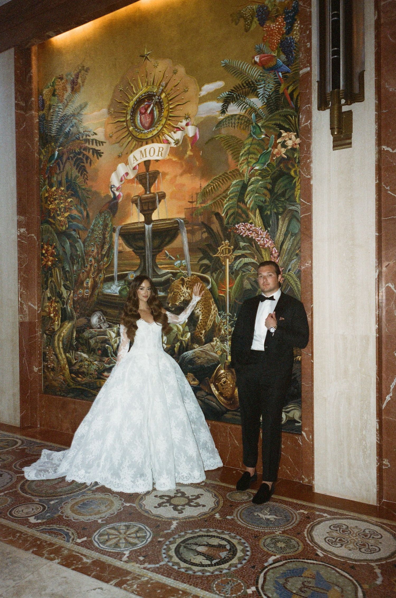 A couple poses in front of an ornate tropical mural with golden sunburst design in an elegant venue setting.