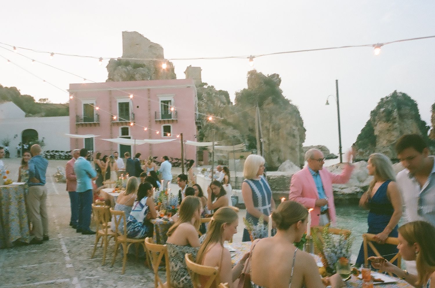 People gather in a small town square during a local celebration with buildings and string lights visible in the background.