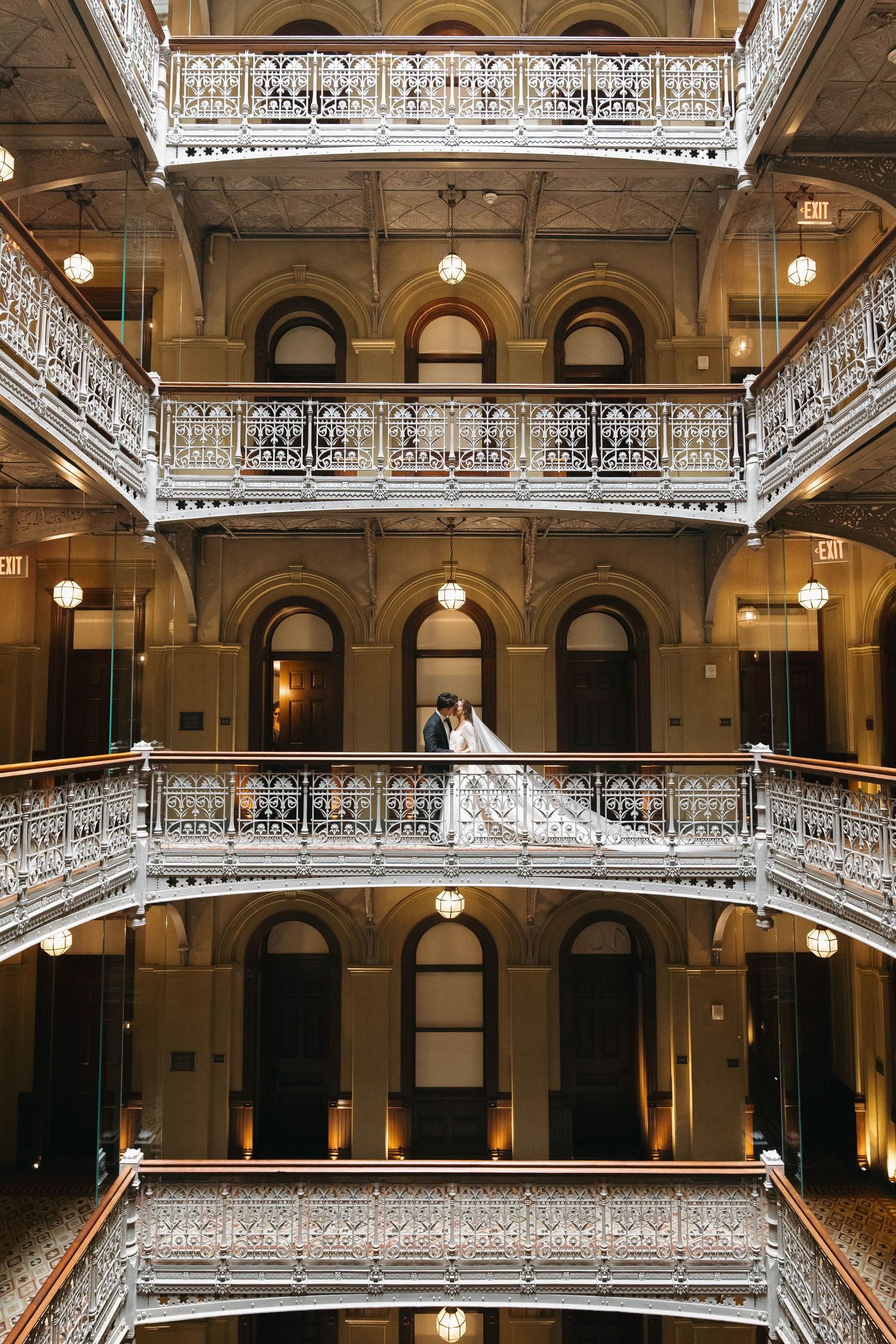 Bride and groom pose on ornate iron balcony inside a grand multi-story atrium with decorative railings.
