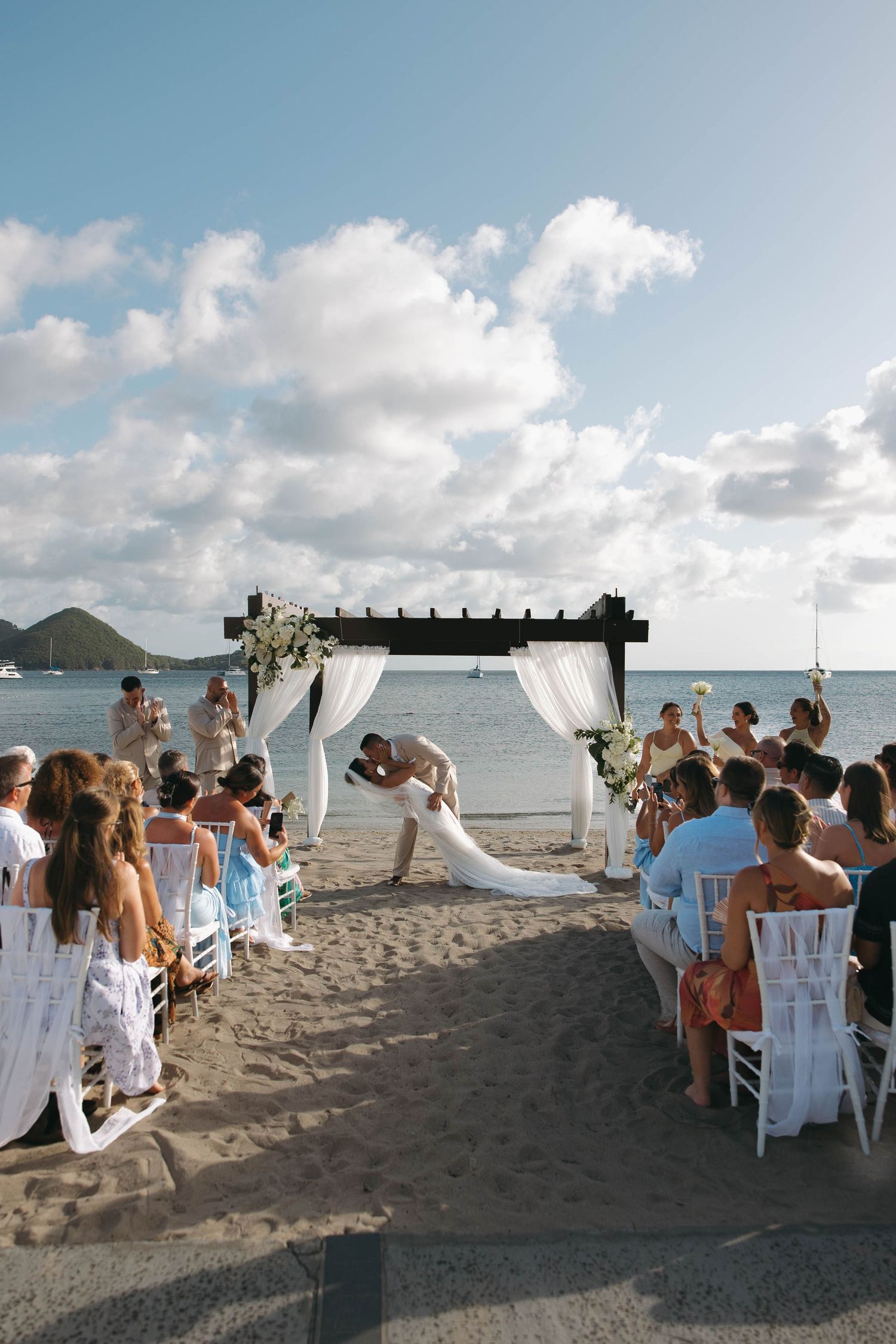 Couple shares first kiss at romantic beach wedding ceremony with white draped arch, ocean backdrop, and seated guests on sandy shore.