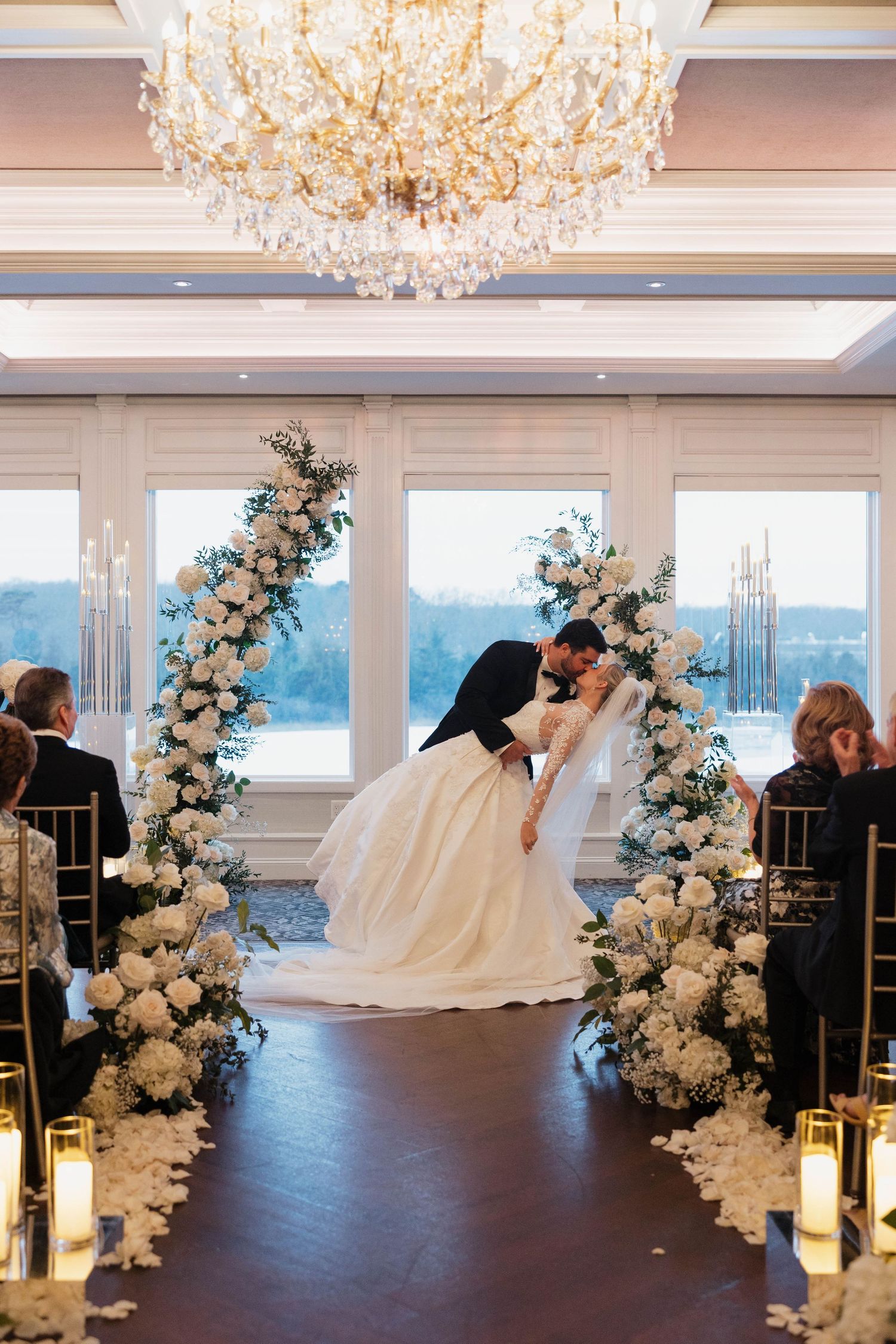 Elegant waterfront wedding ceremony with white floral arches and crystal chandeliers overhead.