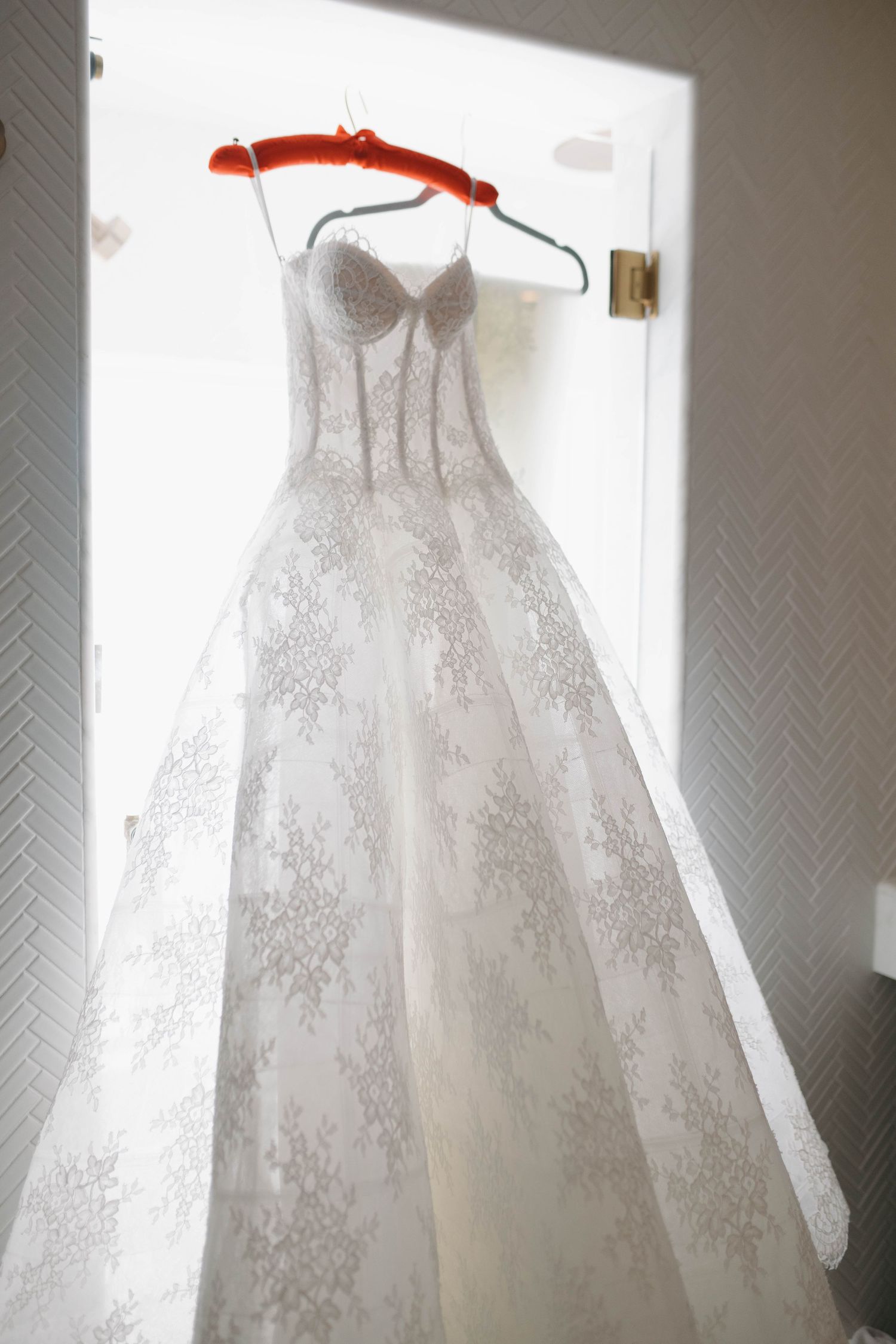 White lace wedding dress with corset bodice hanging against textured wall in natural light.