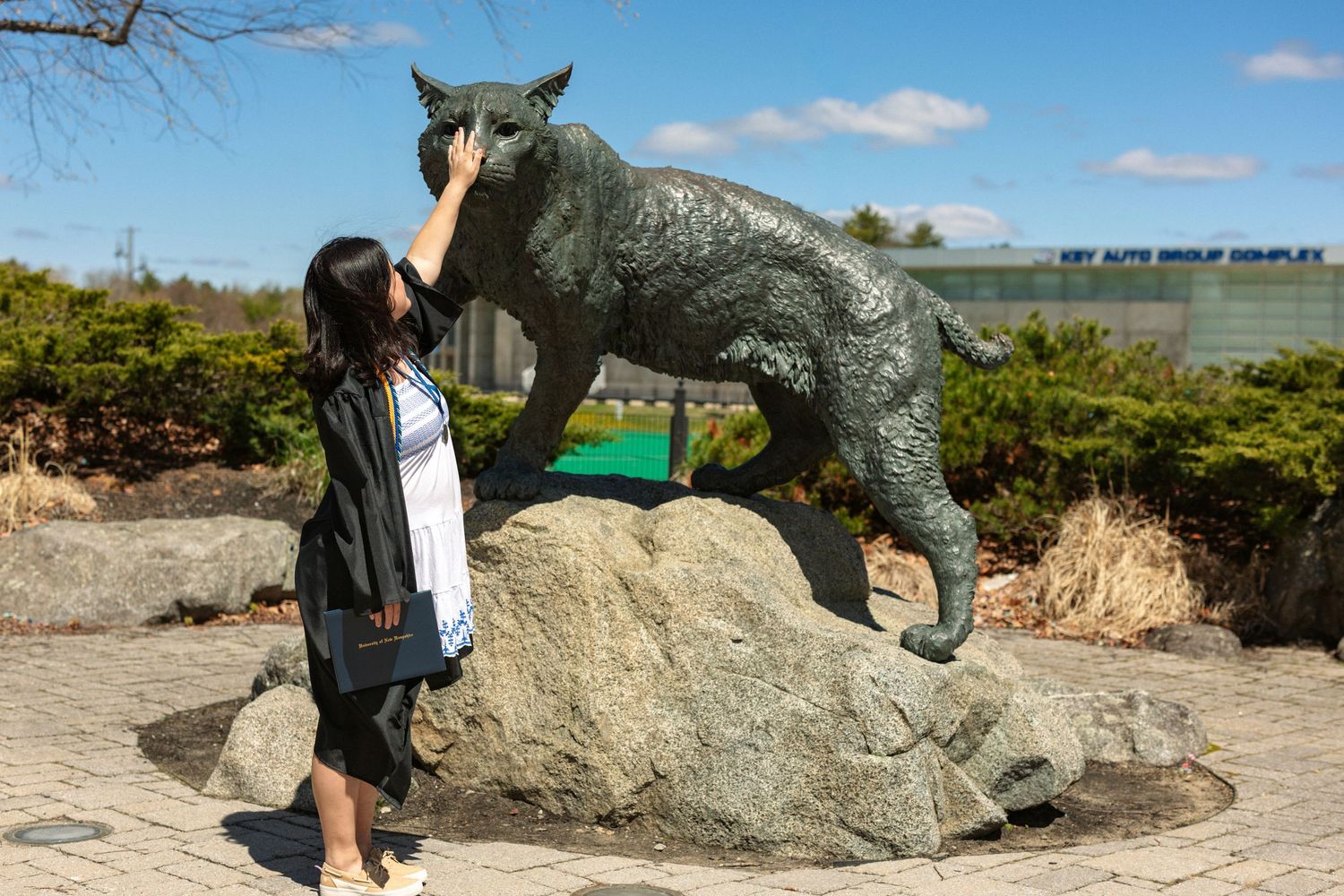 A woman in graduation attire poses touching a bronze wolf statue on a rock outdoors on a sunny day.