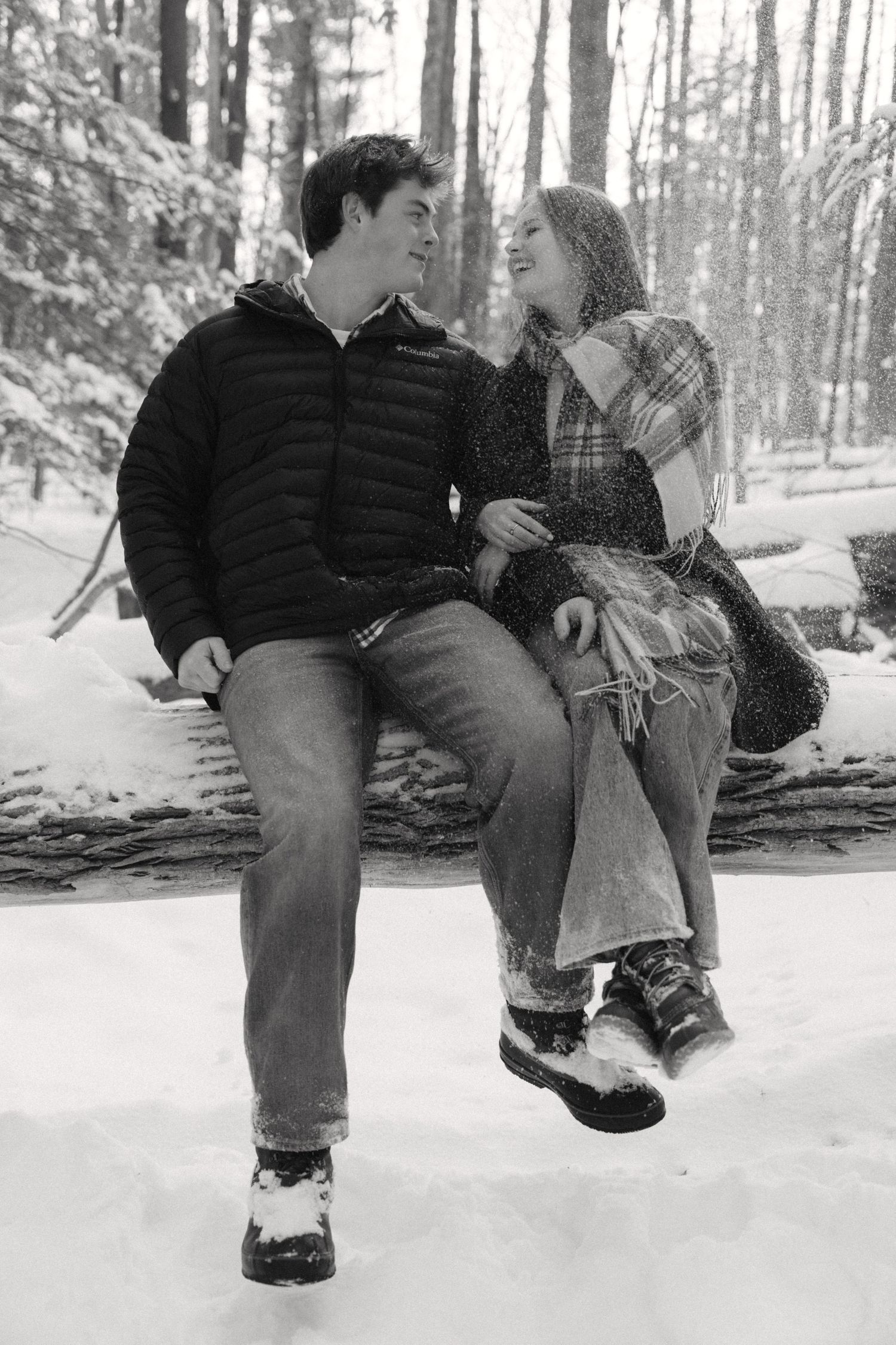 A couple sits together on a fallen log in a snowy winter forest, sharing a romantic moment in black and white.