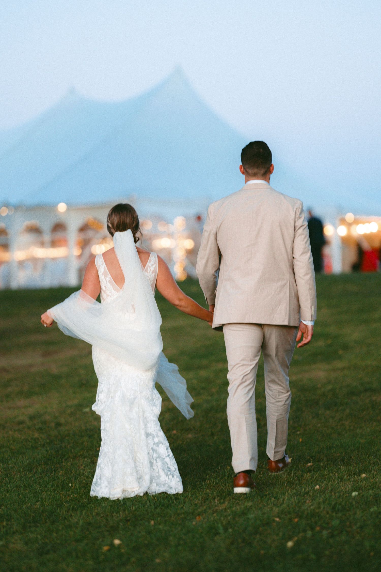 Couple in wedding attire walking together across grassy field at dusk.