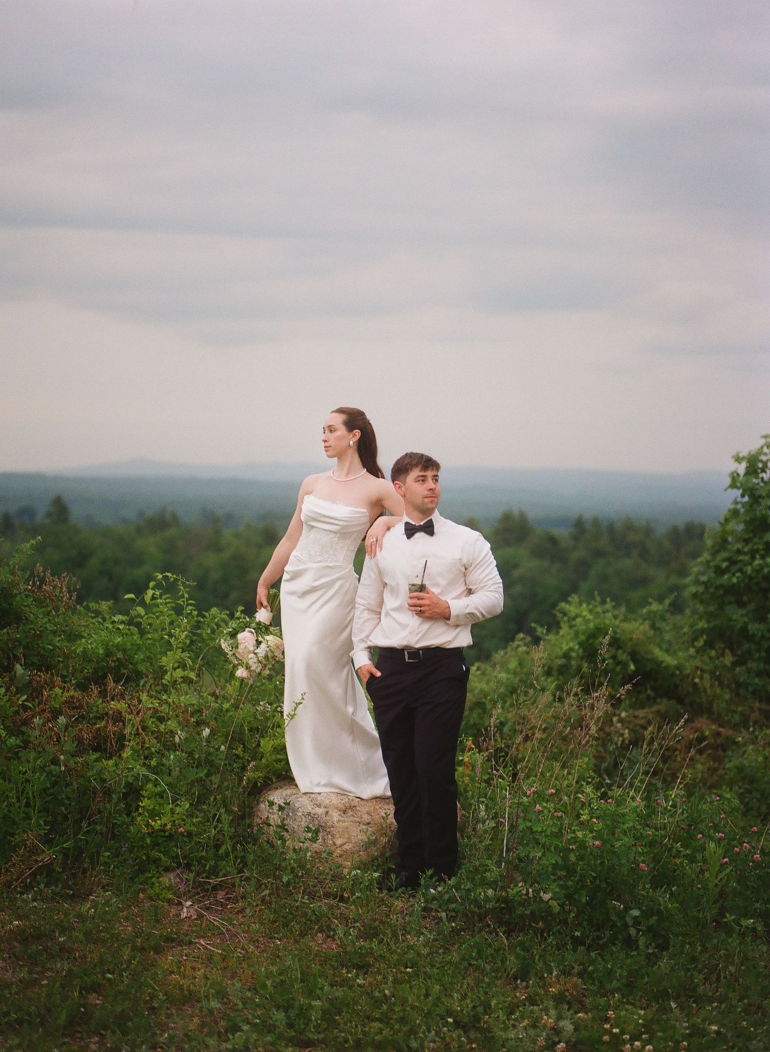 Bride in white gown stands on rock with groom in tuxedo amid lush greenery and rolling hills backdrop.