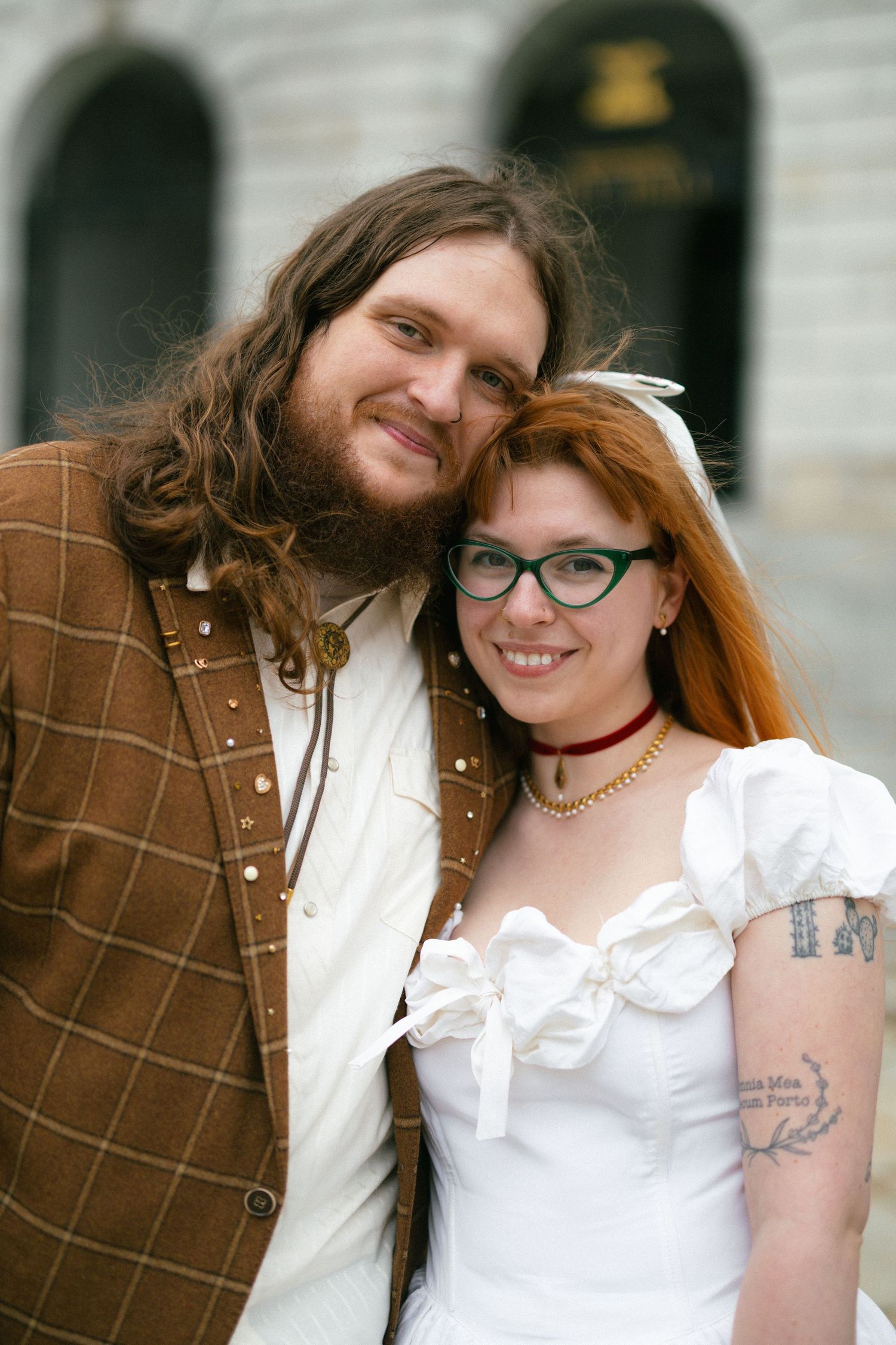 A bearded man in a brown jacket poses with a smiling redhead woman in a white wedding dress and green glasses.