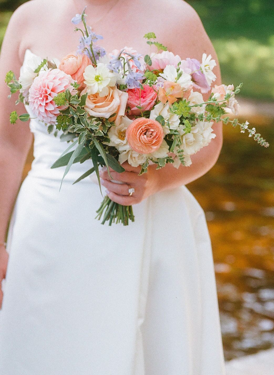 Bride holding a lush peach and pink wedding bouquet with roses, dahlias, and greenery in a white strapless gown.