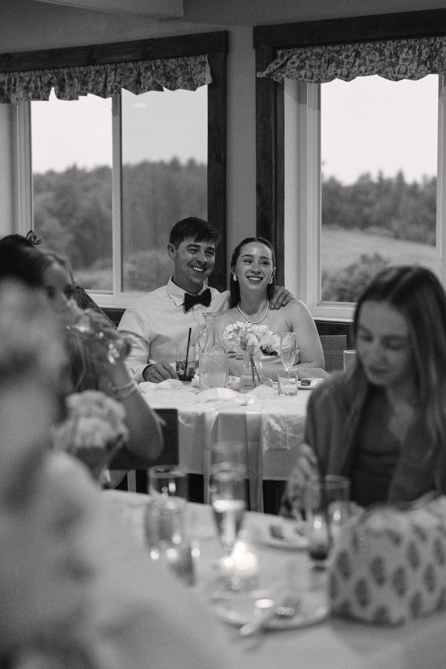 Black and white photo of smiling bride and groom seated at reception table near large windows.