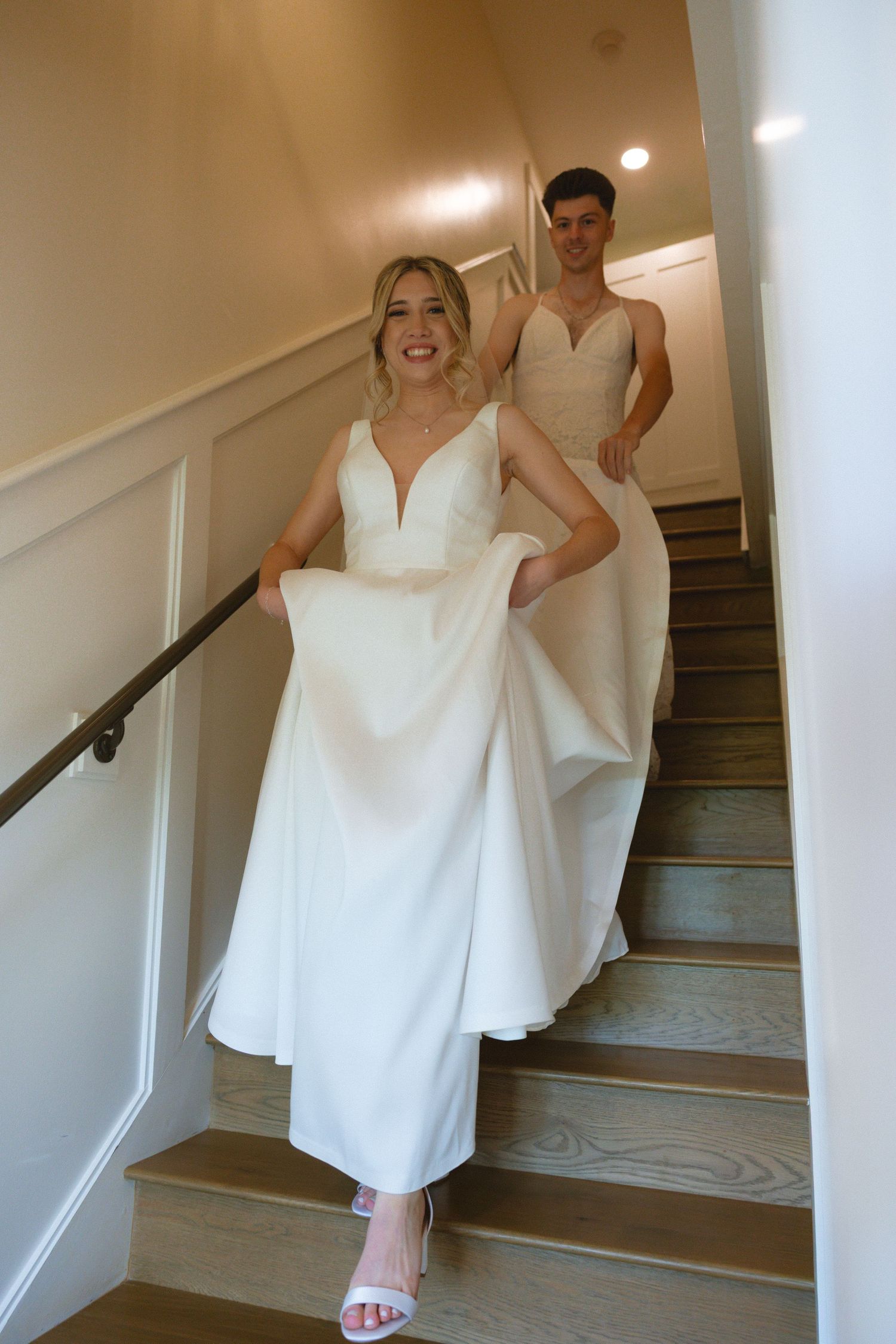 Bride in white gown lifts dress while descending staircase with groom in neutral suit behind her.