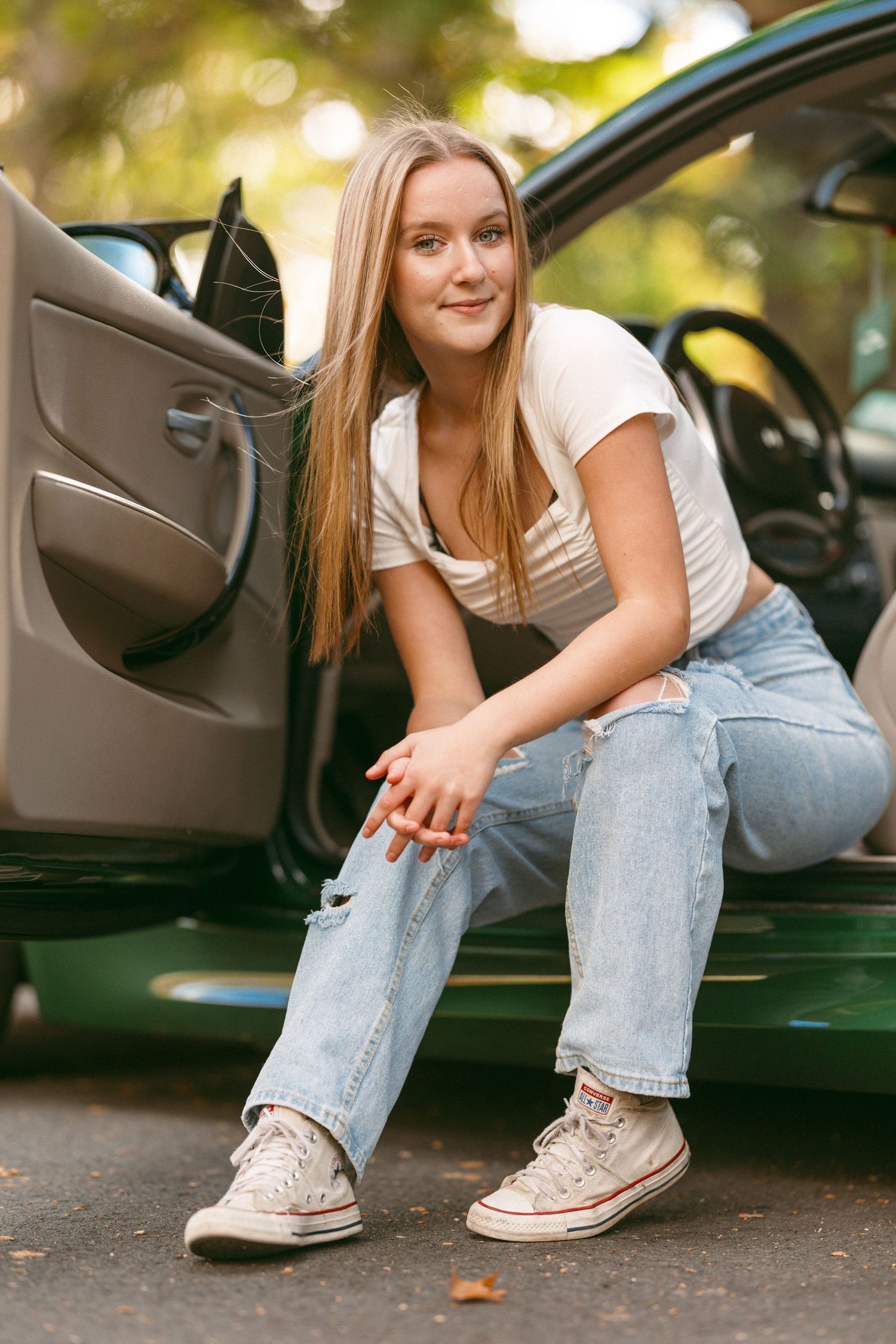 Senior poses by car wearing distressed jeans and white sneakers.