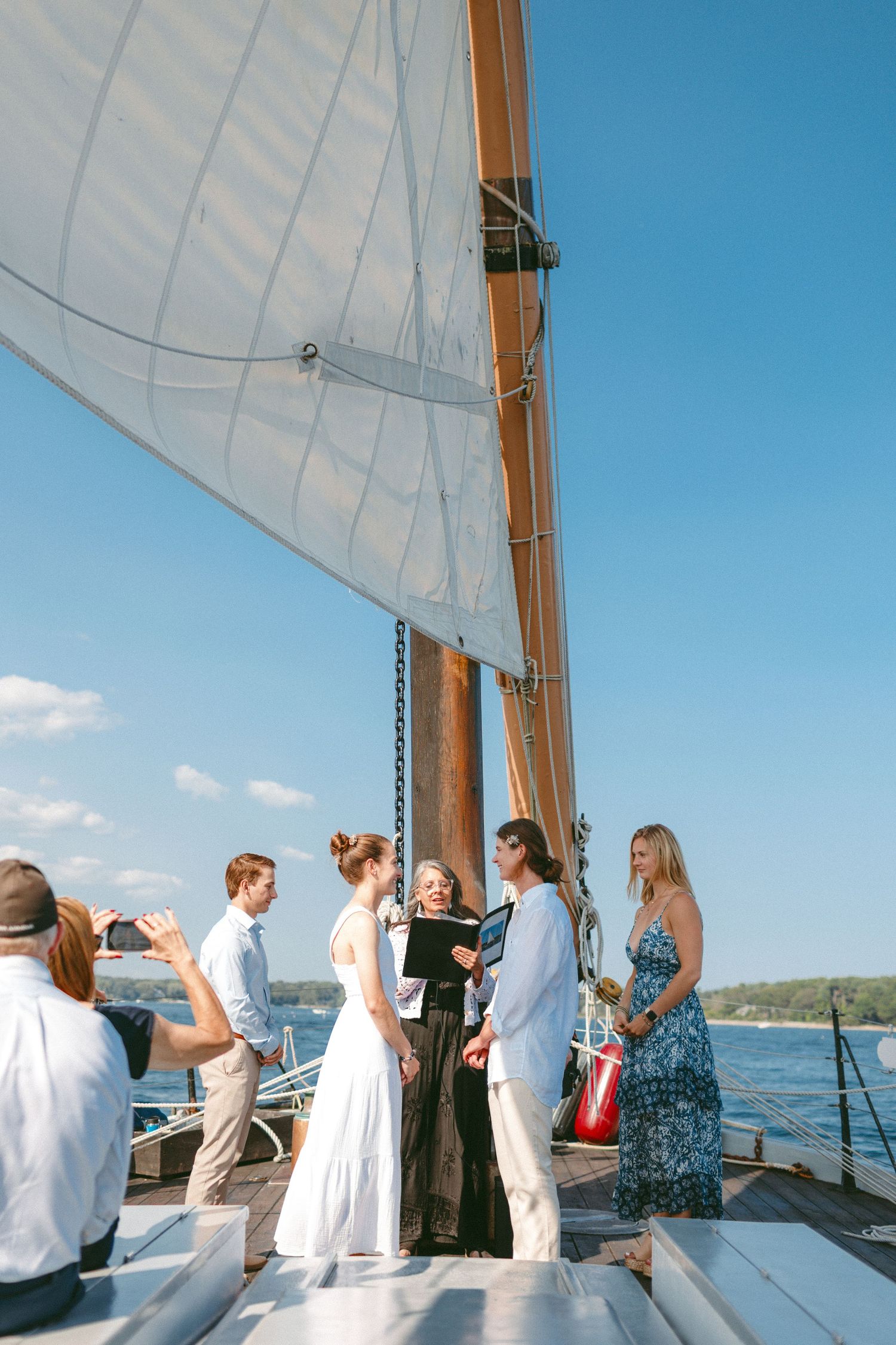 Wedding ceremony in progress on a sailboat deck with white sails and blue sky backdrop.