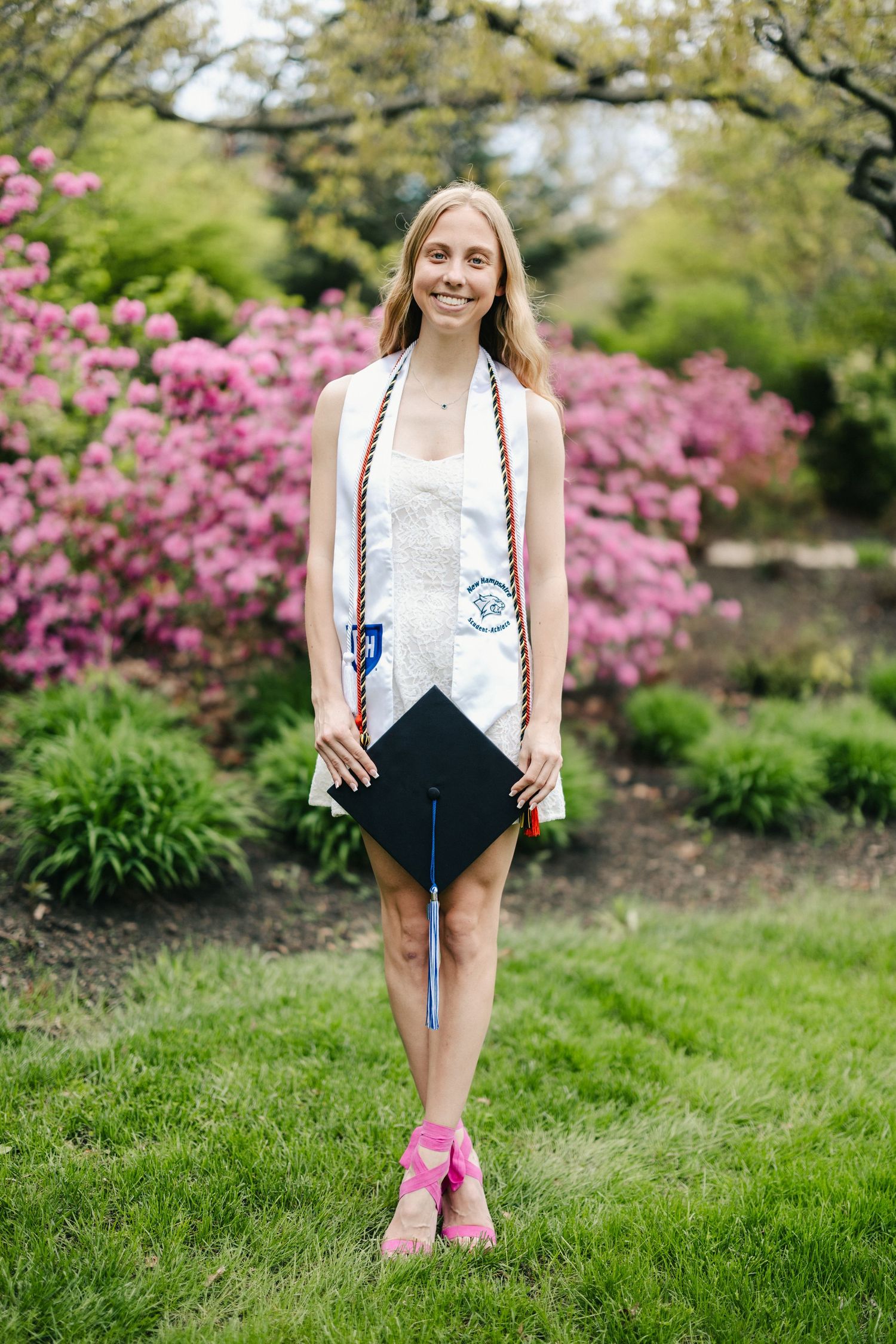 Graduate in white dress poses with graduation cap among blooming pink azalea bushes.