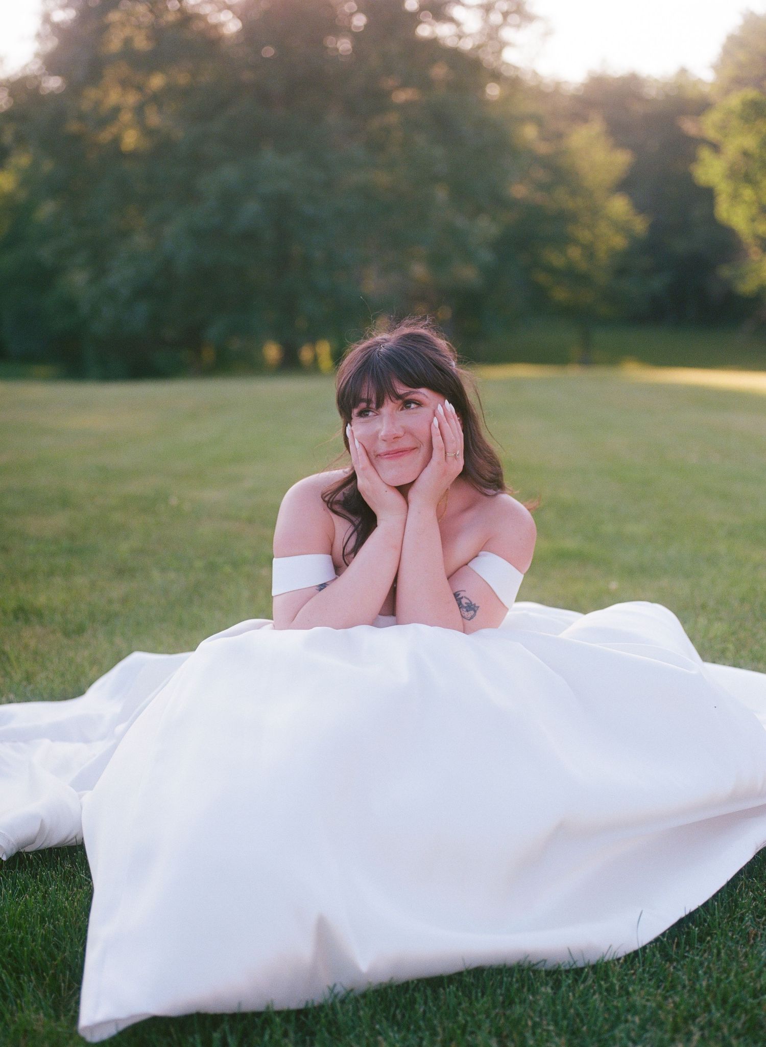 Bride in white off-shoulder gown sitting on green grass, resting chin on hands, smiling dreamily at golden hour.