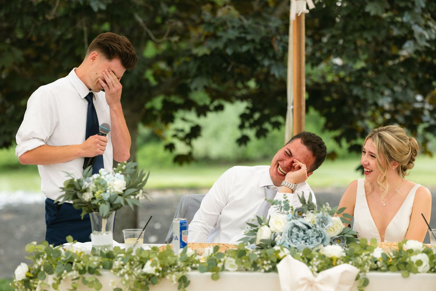 Groom and bride laughing at wedding reception while best man gives emotional speech at outdoor ceremony.