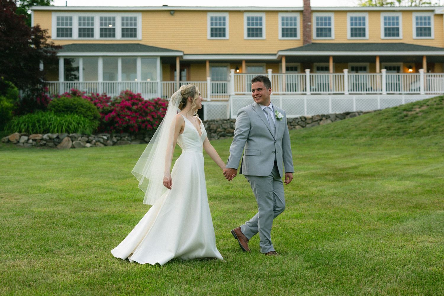 Bride and groom walk hand in hand on green lawn before a large yellow wedding venue.