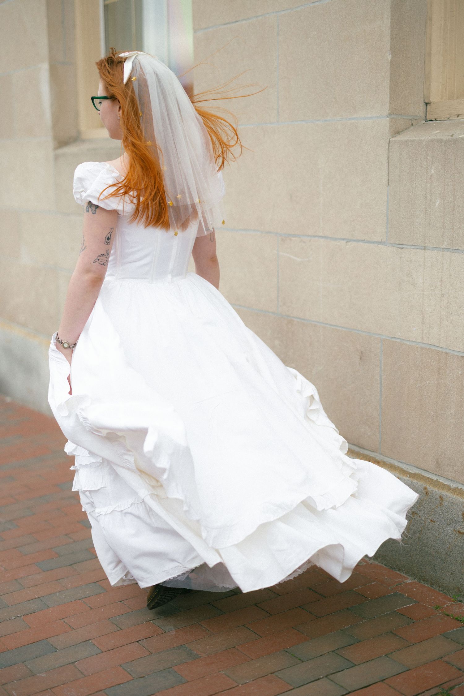 Red-haired bride in white wedding gown and veil walks along brick sidewalk beside stone building.