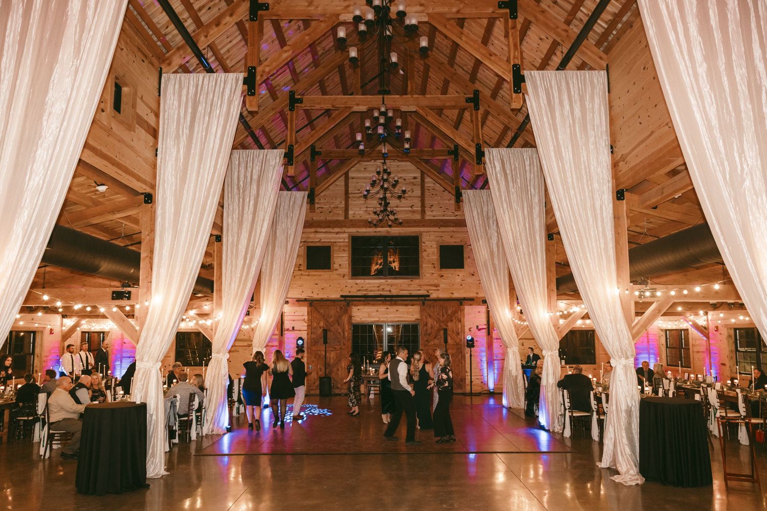 A panoramic view of an elegant ballroom with white drapes, chandeliers and decorative lighting for a wedding reception.