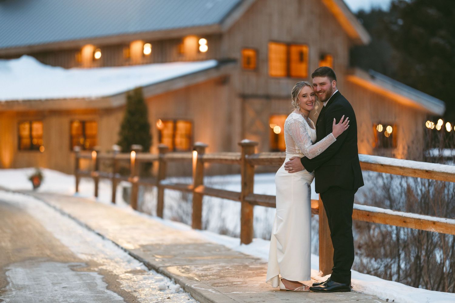 Couple standing together on a snowy deck at dusk with warm glowing lights at a winter wedding venue.