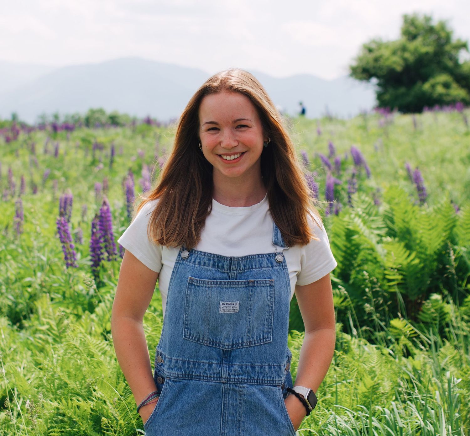 A person wearing denim overalls stands smiling in a field of purple lupine wildflowers on a sunny summer day.