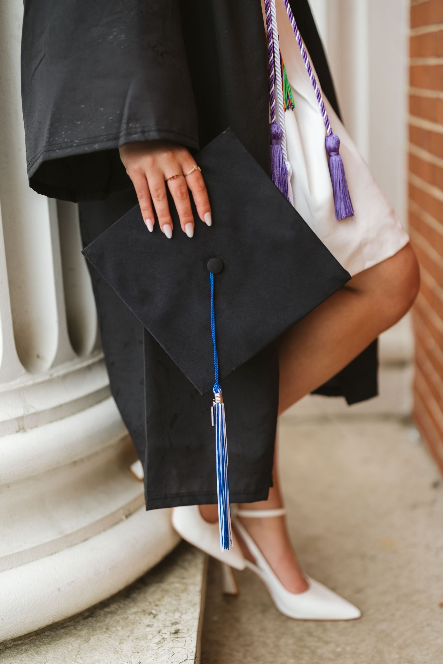 Graduate in black gown holds mortarboard cap with blue tassel, wearing white heels near decorative columns.