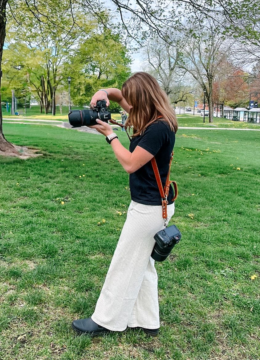Photographer in white dress taking photos in a grassy park setting.