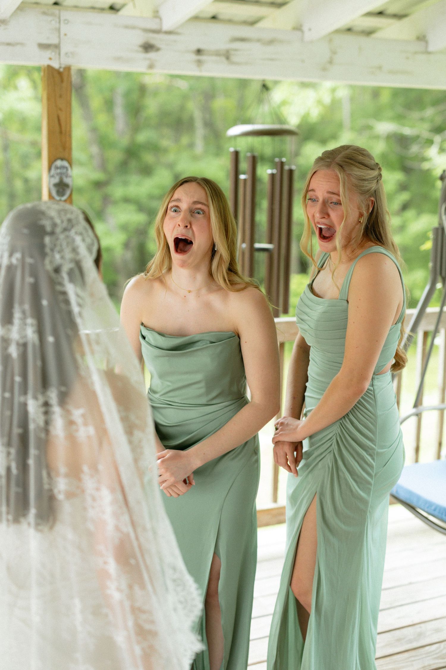 Two bridesmaids in sage green dresses reacting with shocked expressions upon seeing the wedding dress.