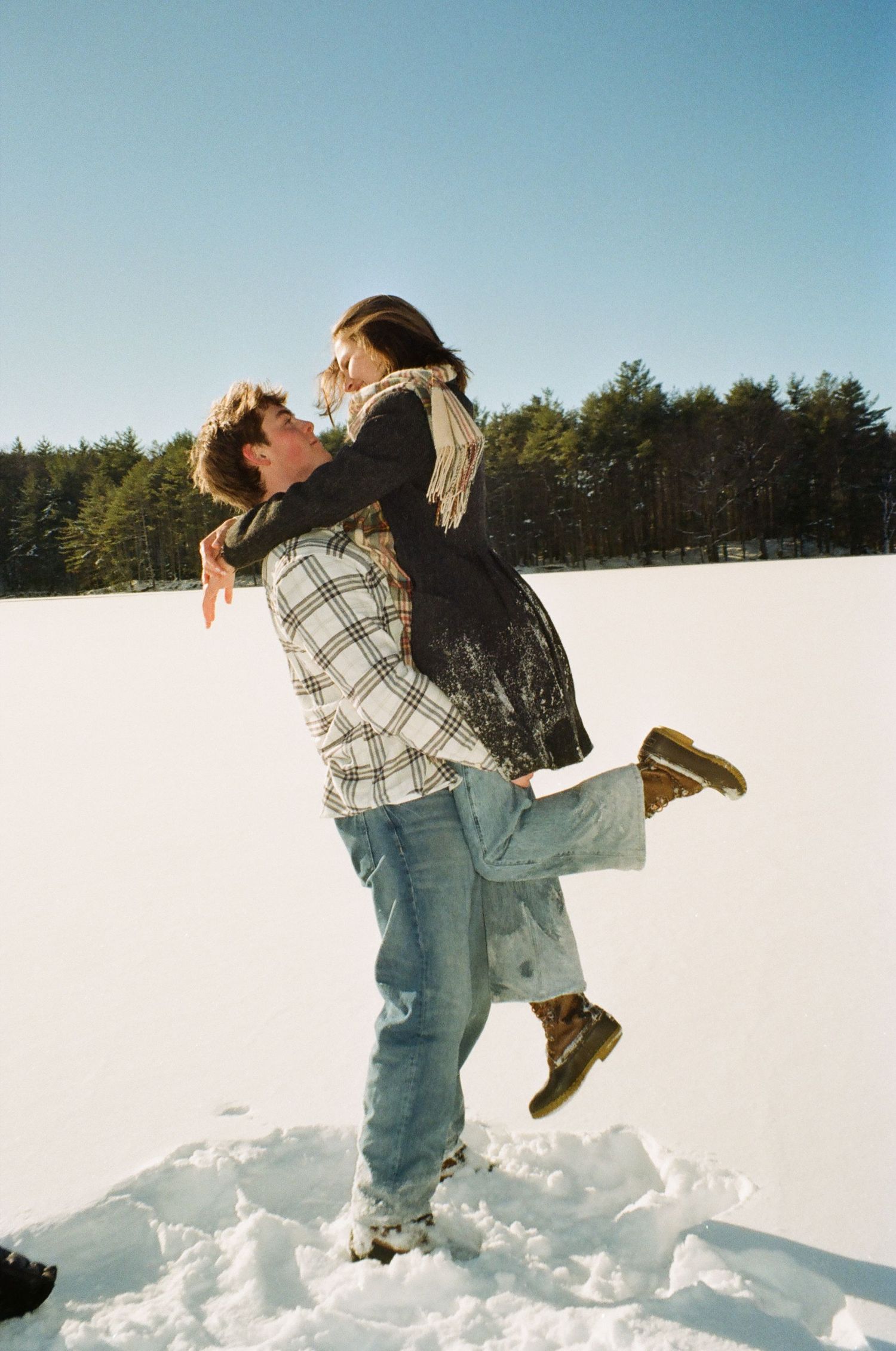 Two people share a romantic embrace while standing in deep snow on a bright winter day with evergreen trees in background.