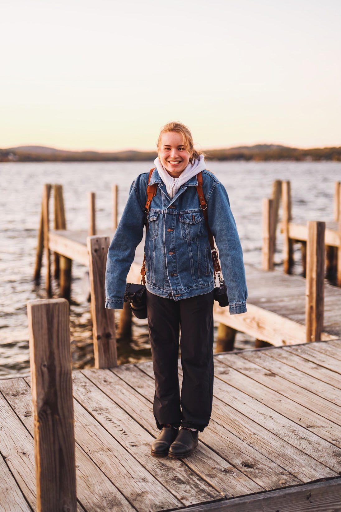 A young child wearing denim stands on a rustic wooden dock overlooking a serene lake at sunset.