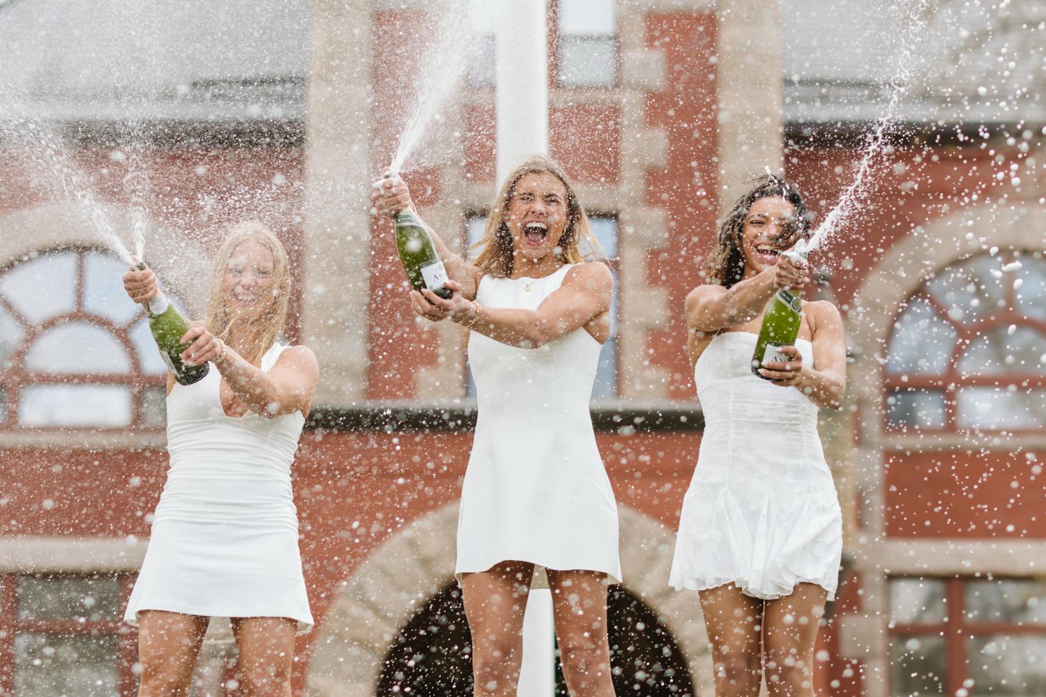 Three women in white dresses celebrate by spraying champagne outdoors in front of a red brick building.