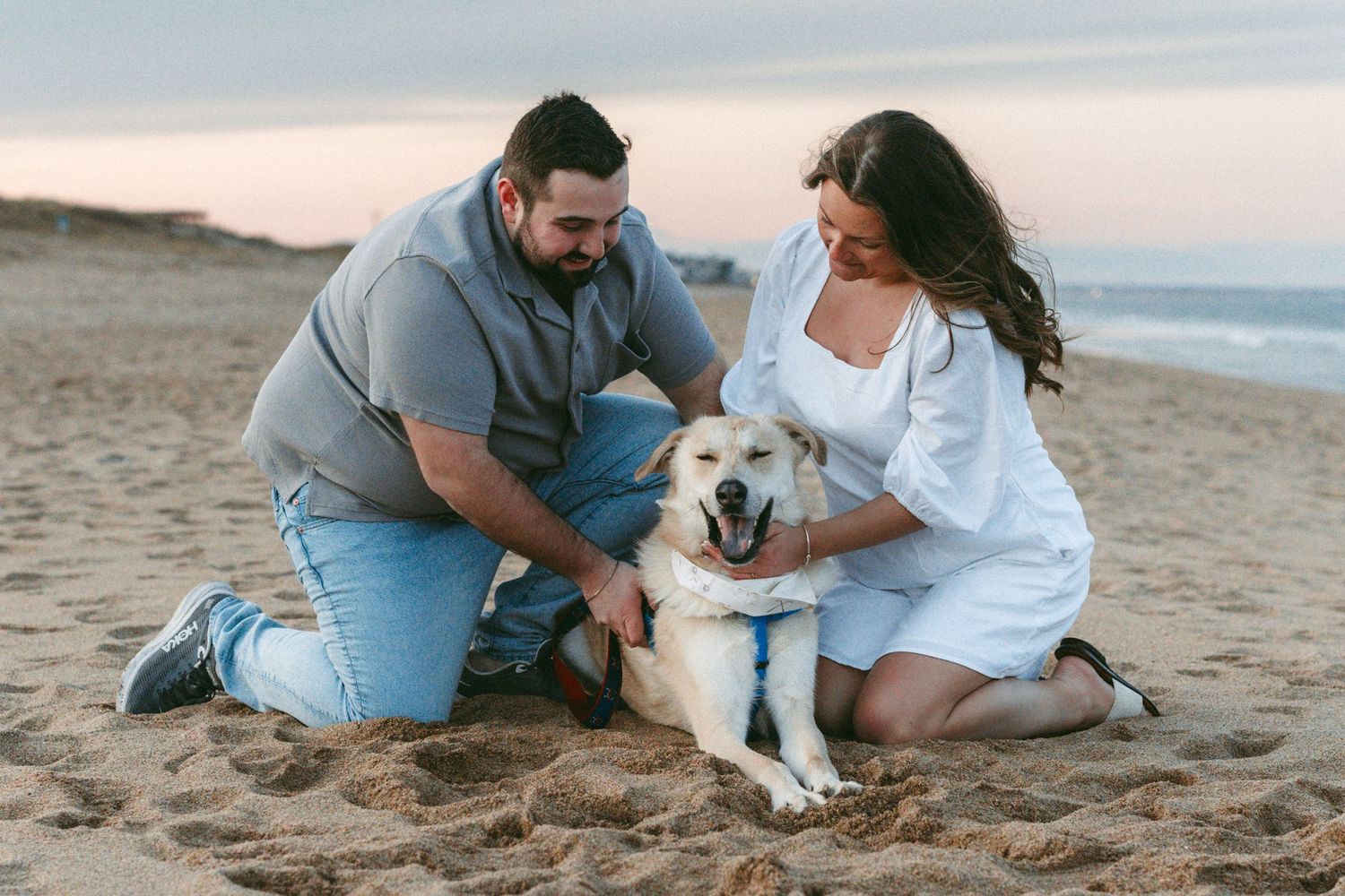 Couple kneeling on sandy beach petting a happy yellow Labrador dog at sunset.