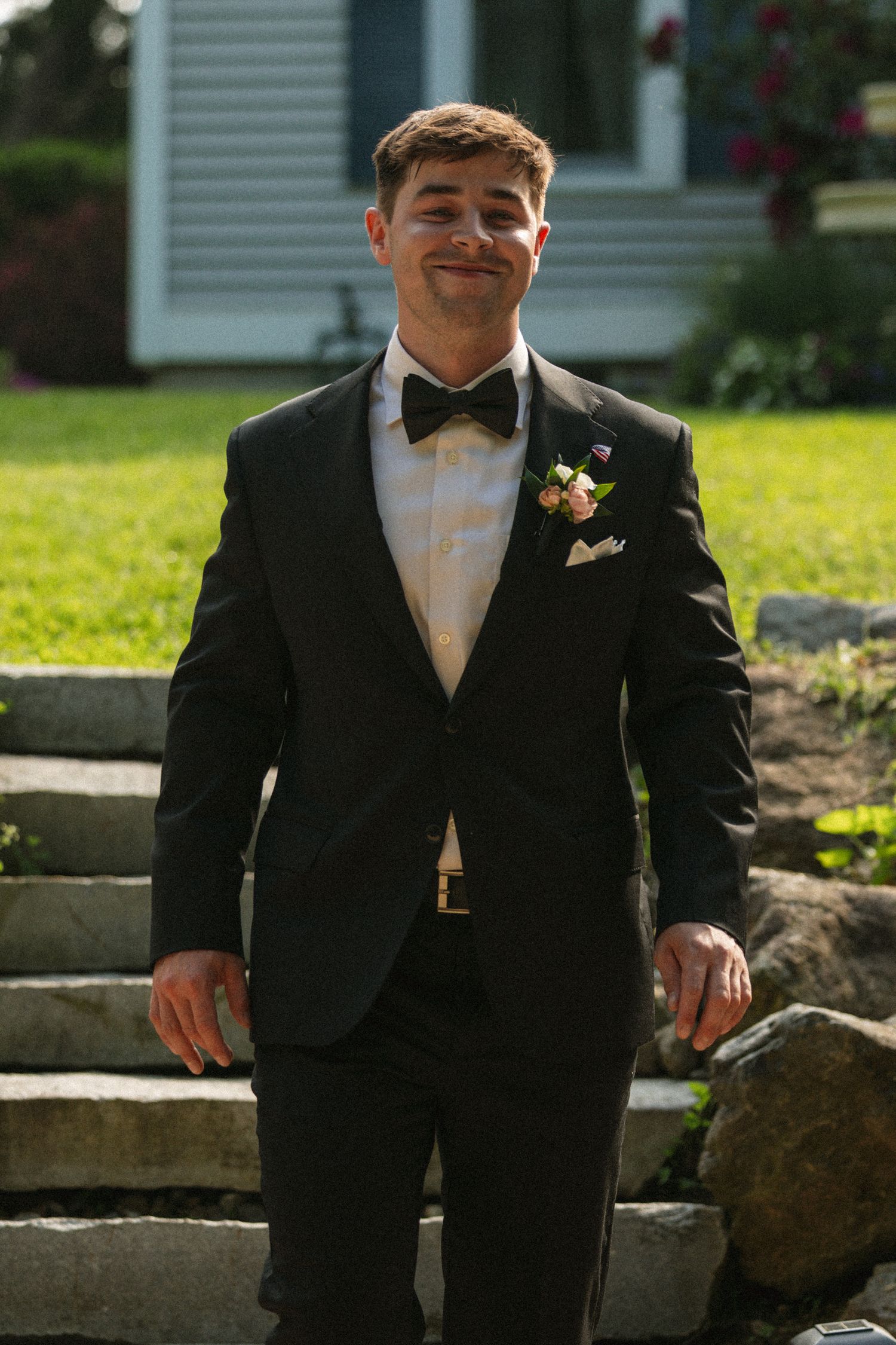 Young man in black tuxedo with bow tie and boutonniere standing on stone steps outdoors.
