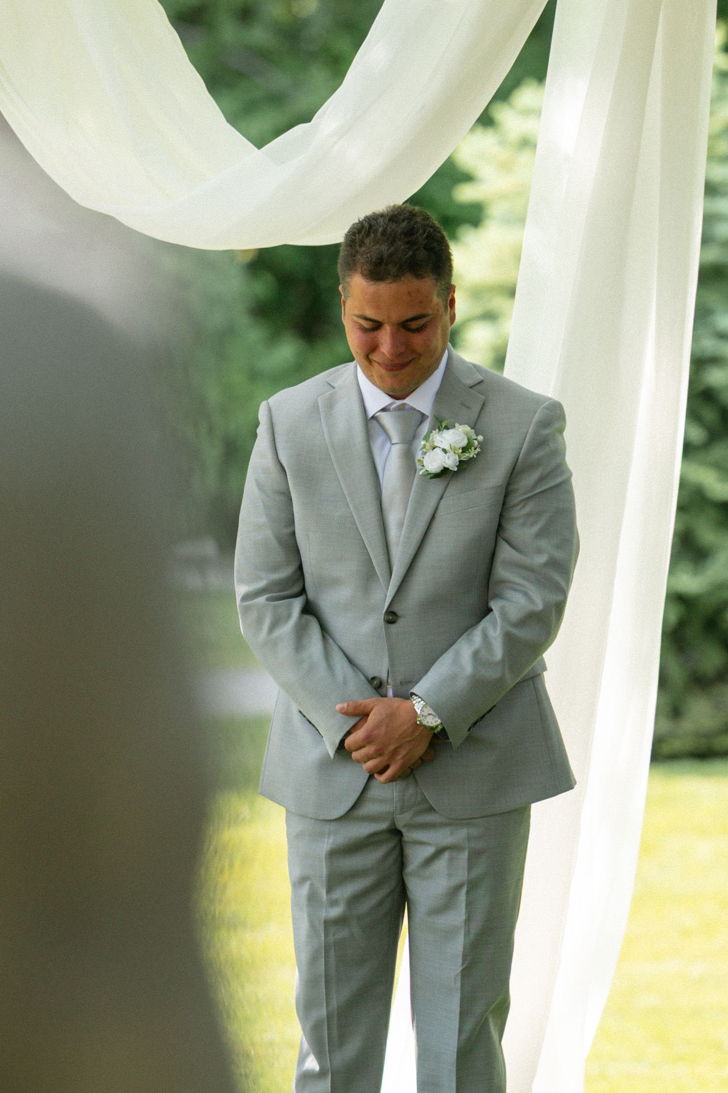 Emotional groom in light gray suit with white boutonniere stands at outdoor wedding ceremony under white draped fabric.