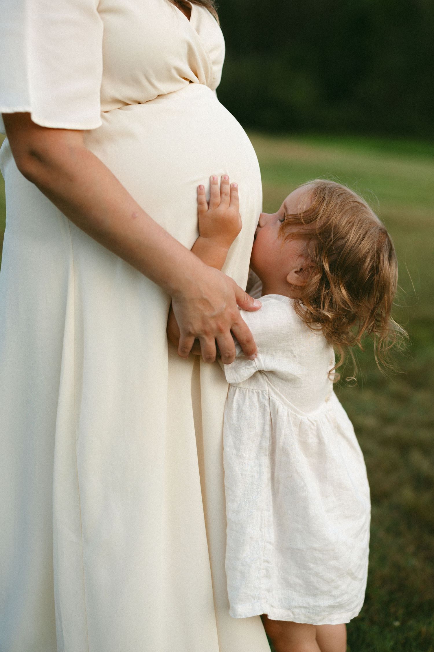 A young girl in a white dress kisses a pregnant woman's belly outdoors on green grass.