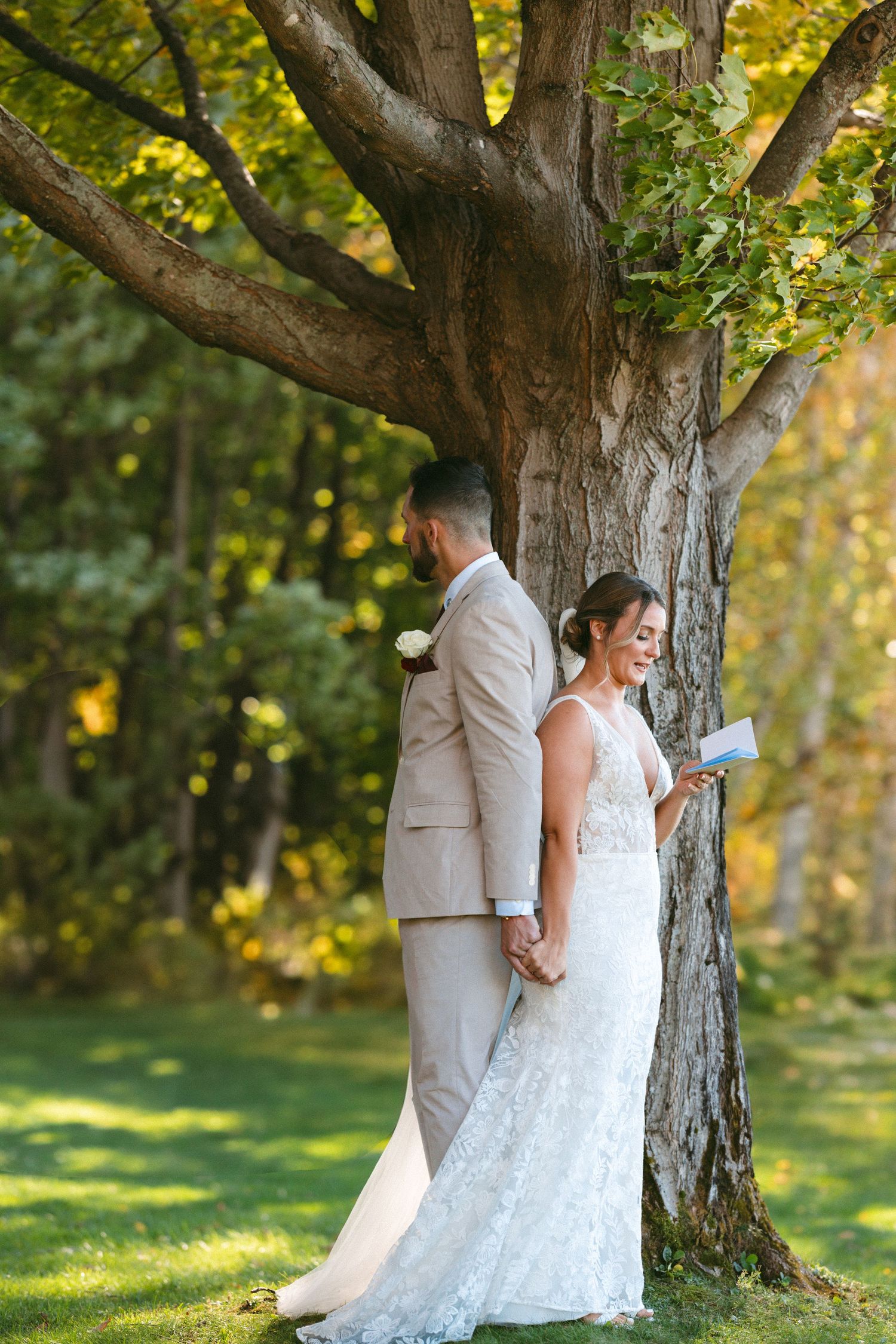 A couple in wedding attire share an intimate moment against a tree trunk with sunlit yellow foliage in the background.