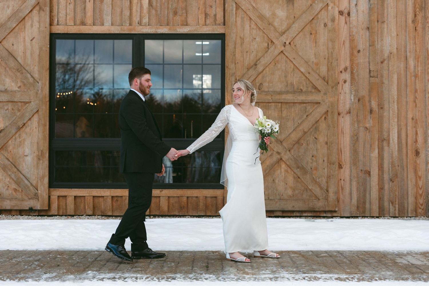A winter wedding scene with a couple holding hands outside a rustic wooden barn in the snow.
