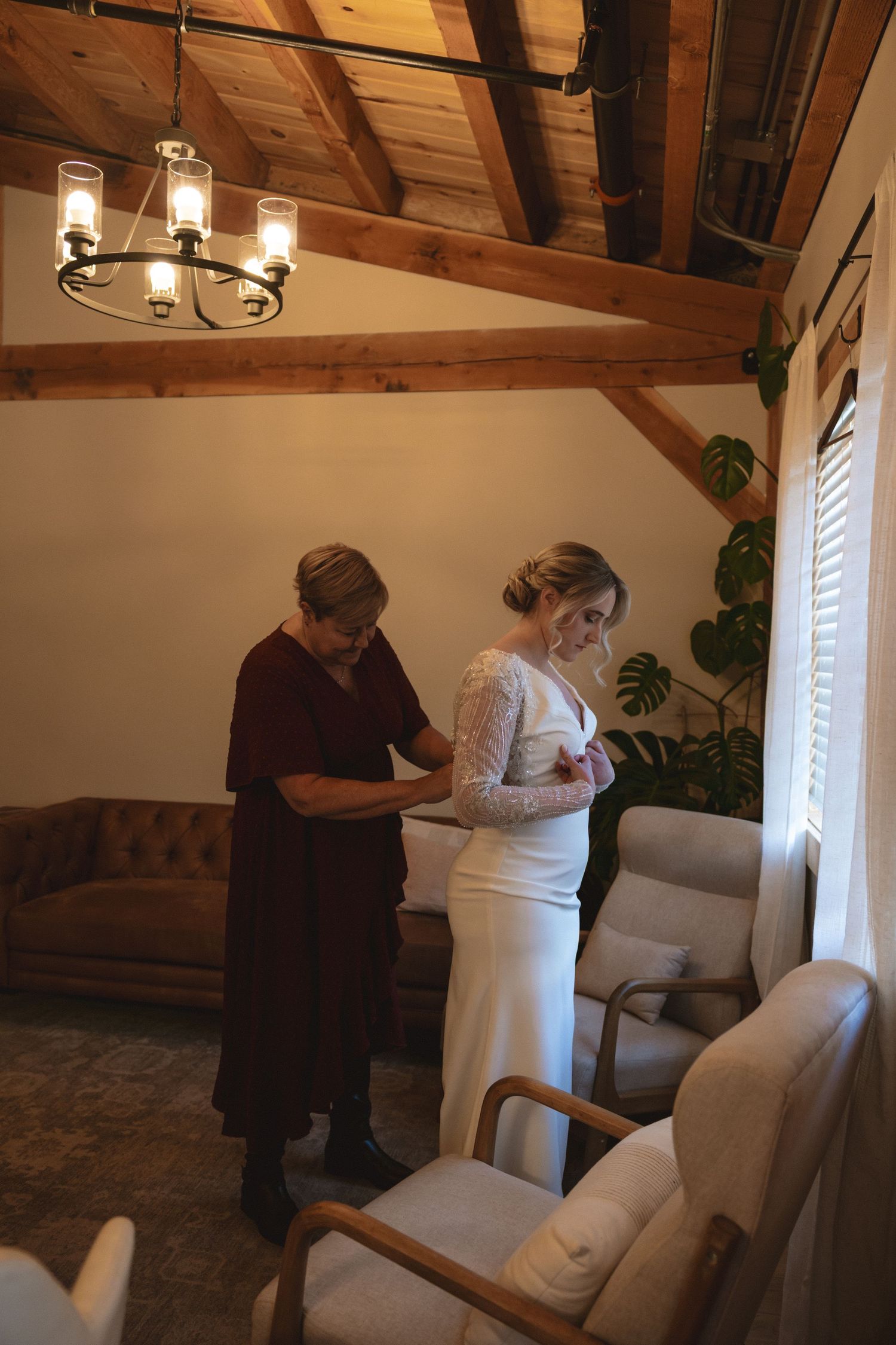 Two people in a rustic room with wooden beams prepare for a special occasion near a window with natural light.