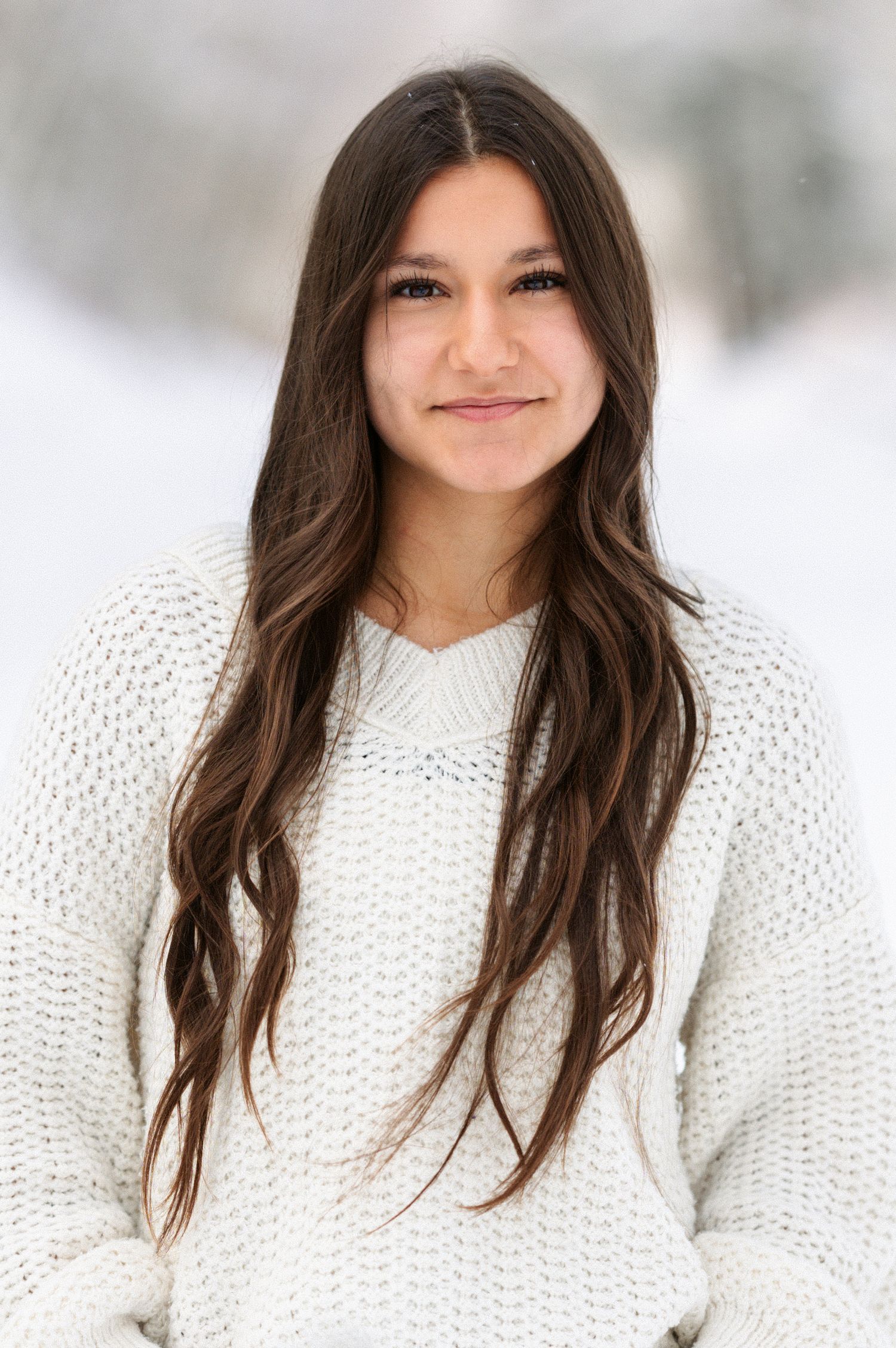 Person wearing a cozy white knit sweater poses in a snowy winter setting with natural lighting and soft background blur.