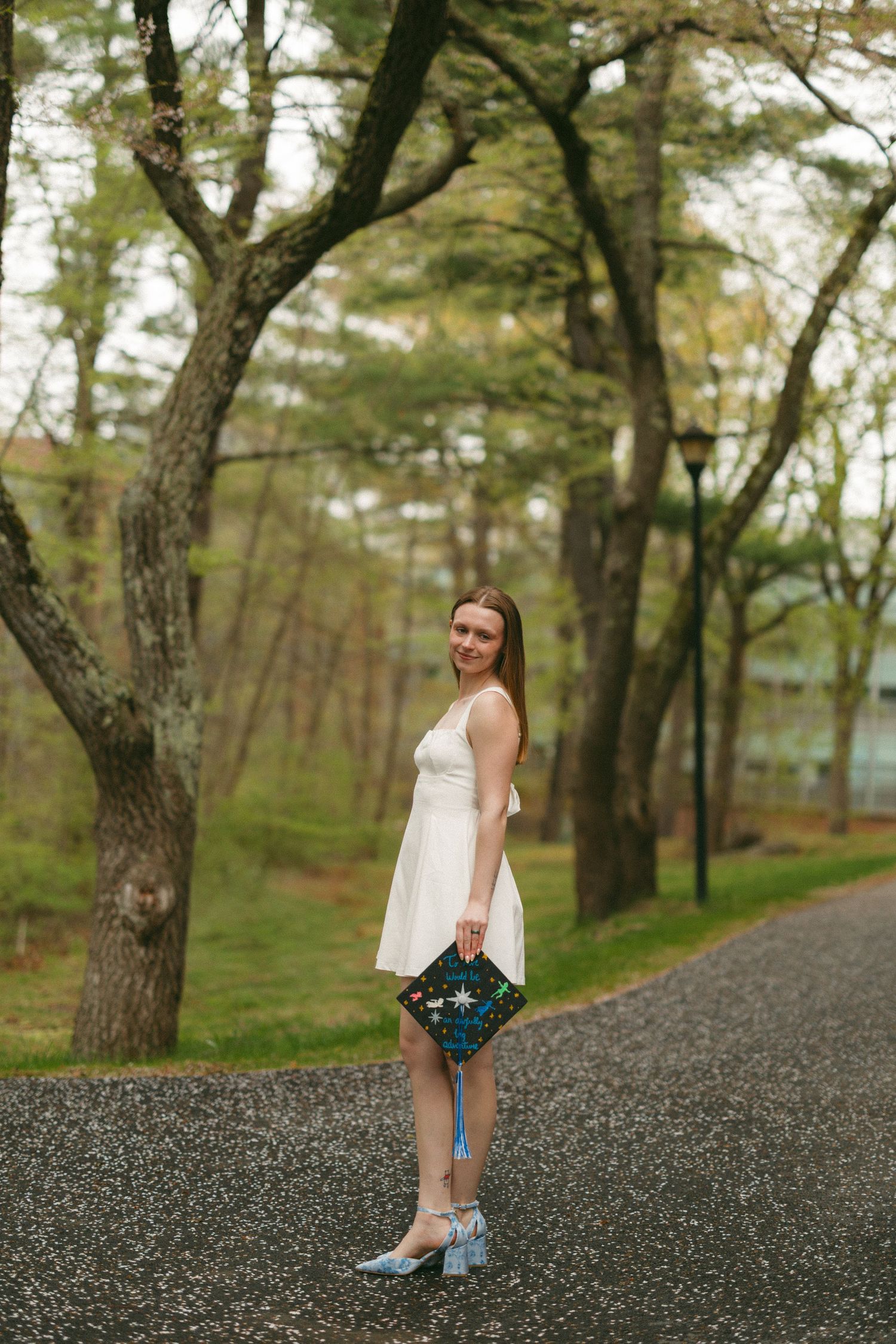 Young woman in white dress holding colorful clutch purse, standing on tree-lined path in lush green park.