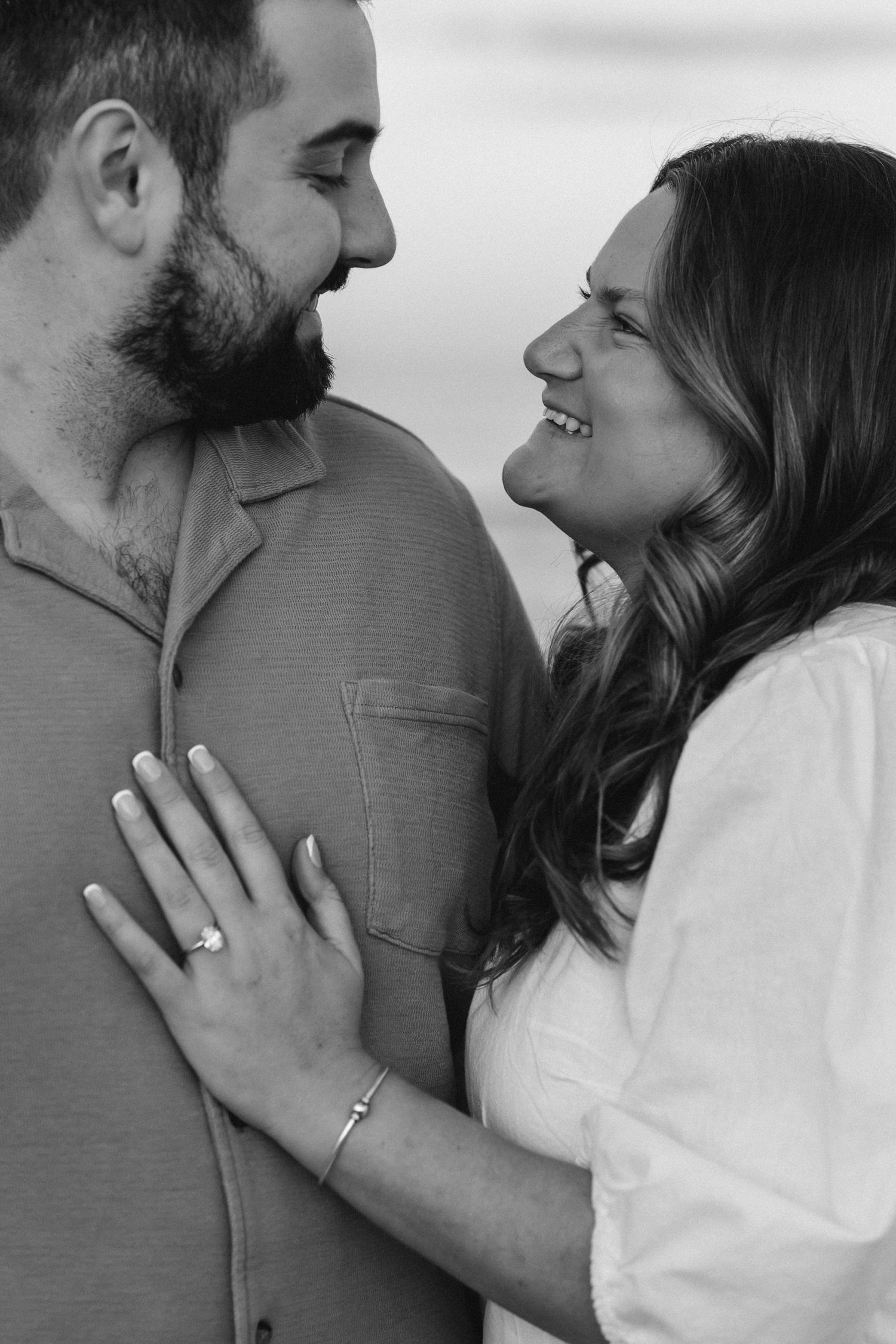 Black and white photo of a smiling couple gazing at each other, woman wearing engagement ring resting hand on mans chest.