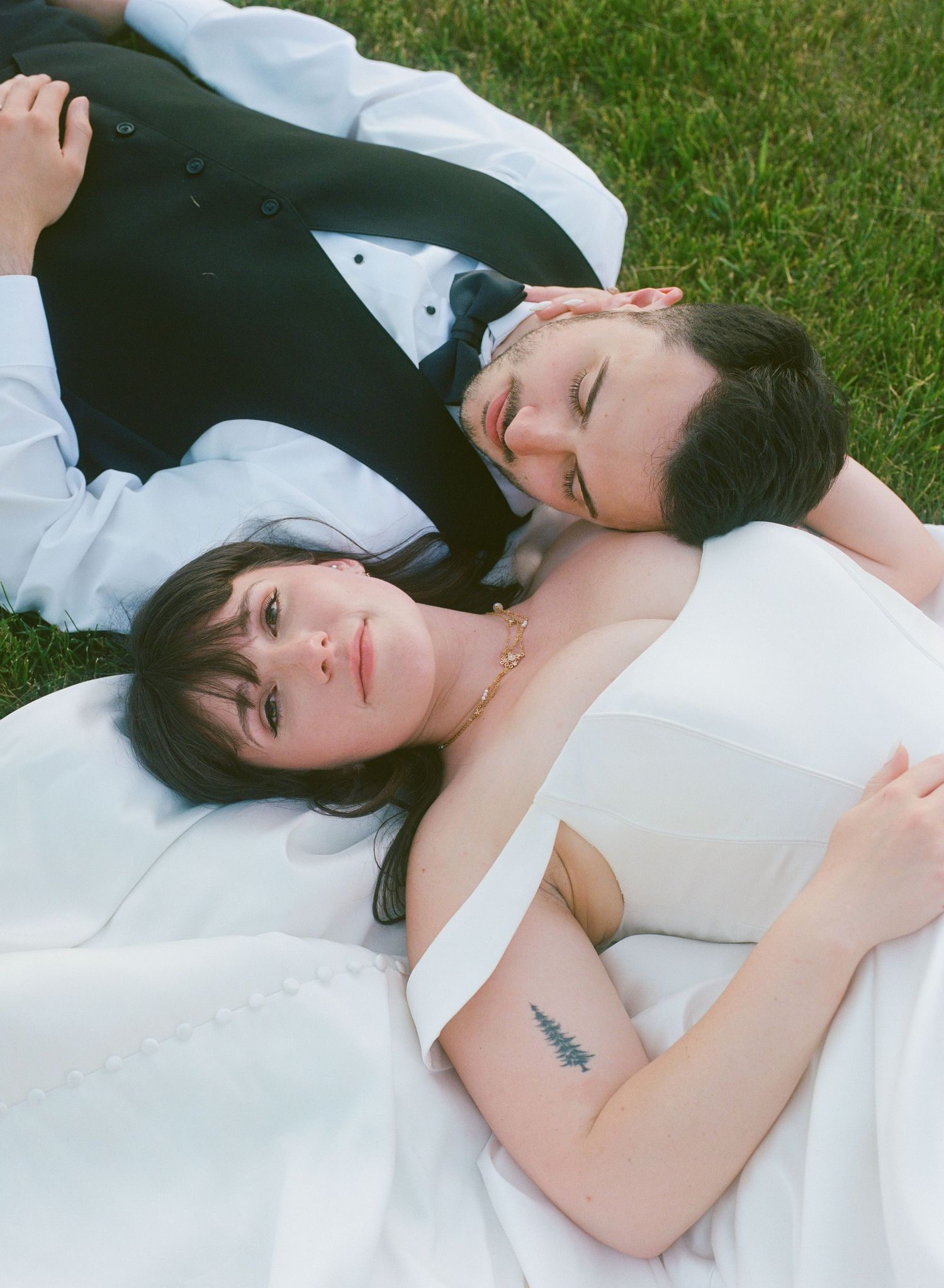 Bride and groom lying on green grass in wedding attire, sharing a tender moment with eyes closed.