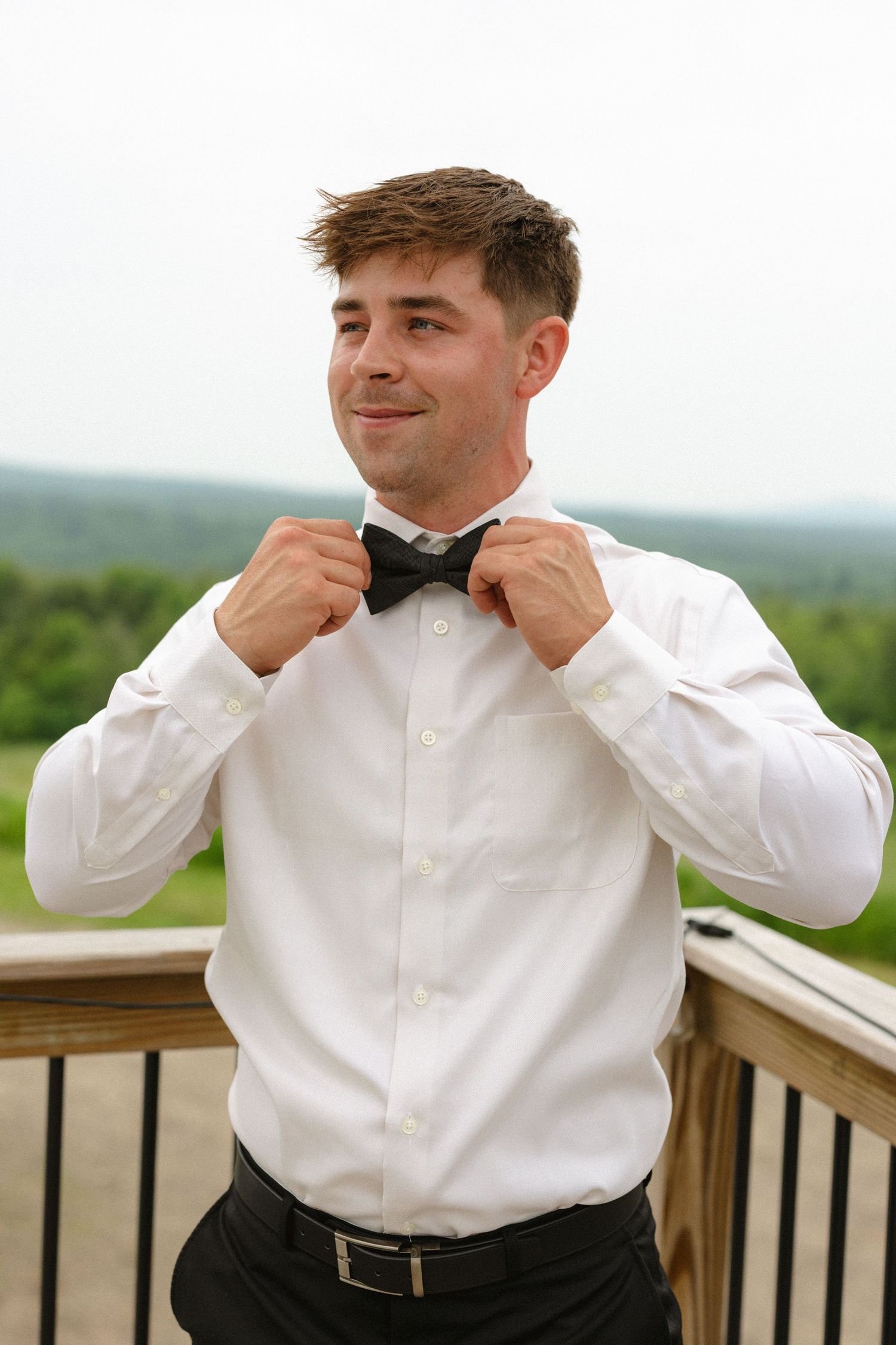 Groom in white dress shirt adjusting black bow tie on wooden deck with green hills in background.