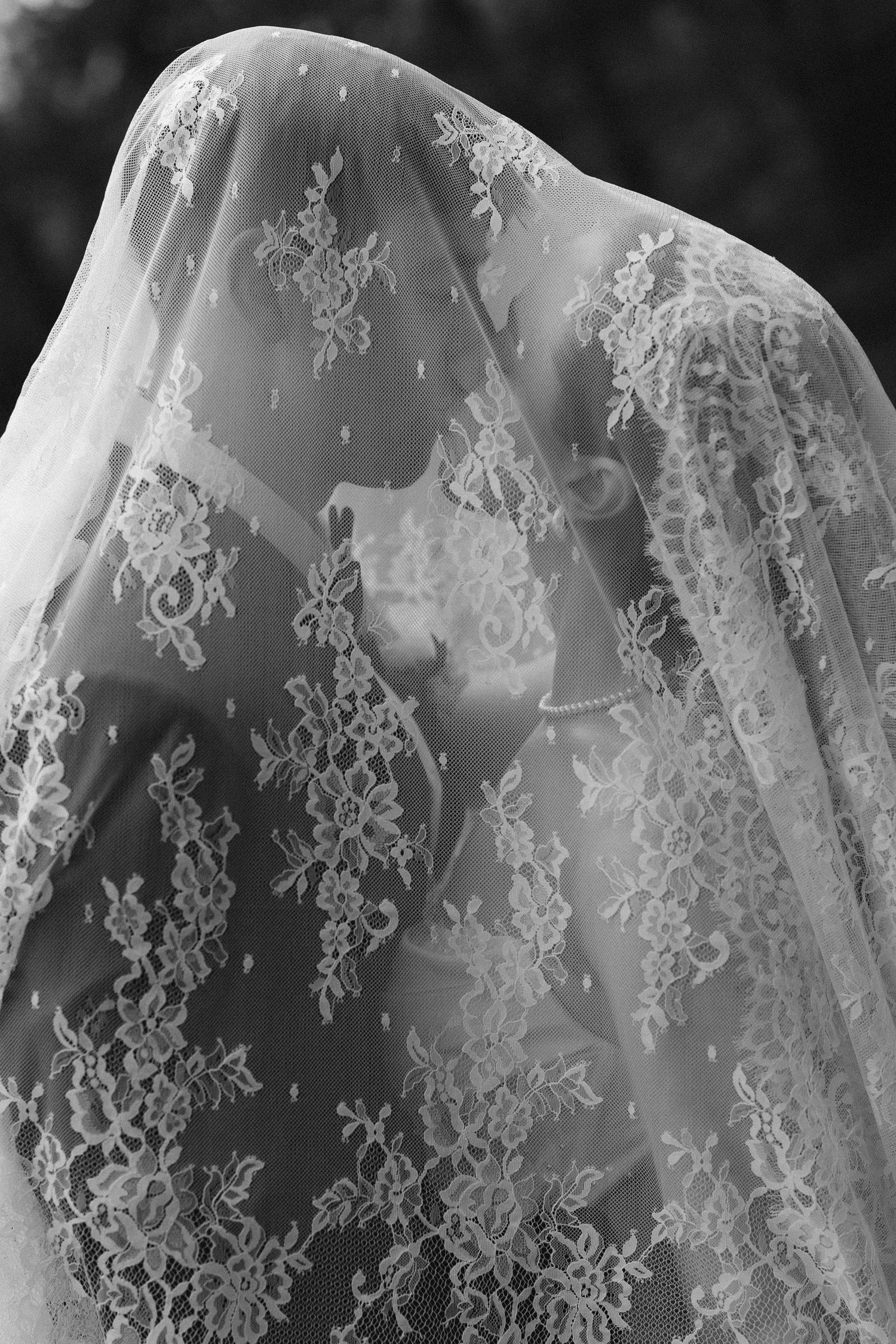 Black and white artistic photo of bride kissing through intricate floral lace wedding veil.