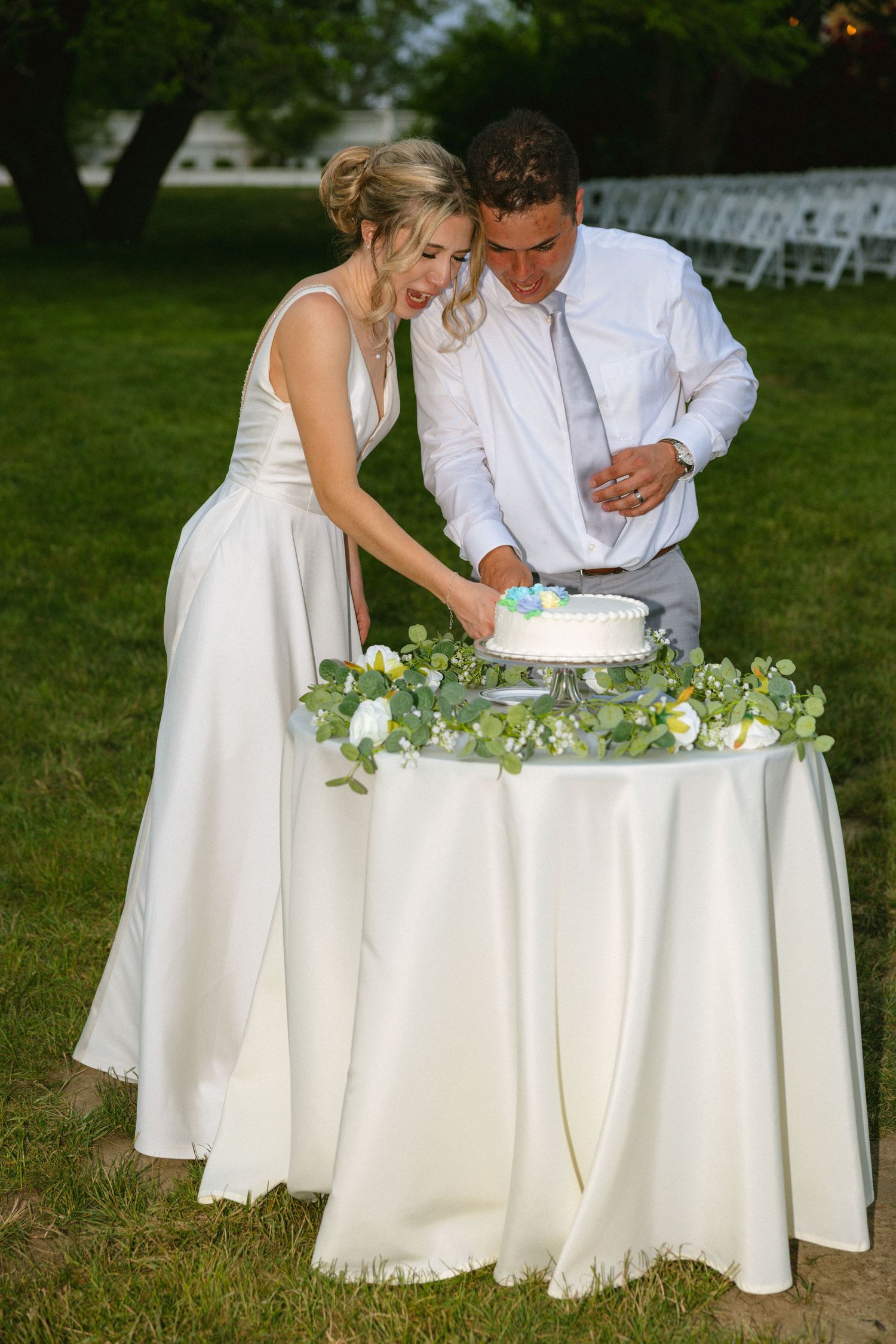 Bride and groom cutting their white wedding cake outdoors on a flower-adorned table during an evening garden reception.
