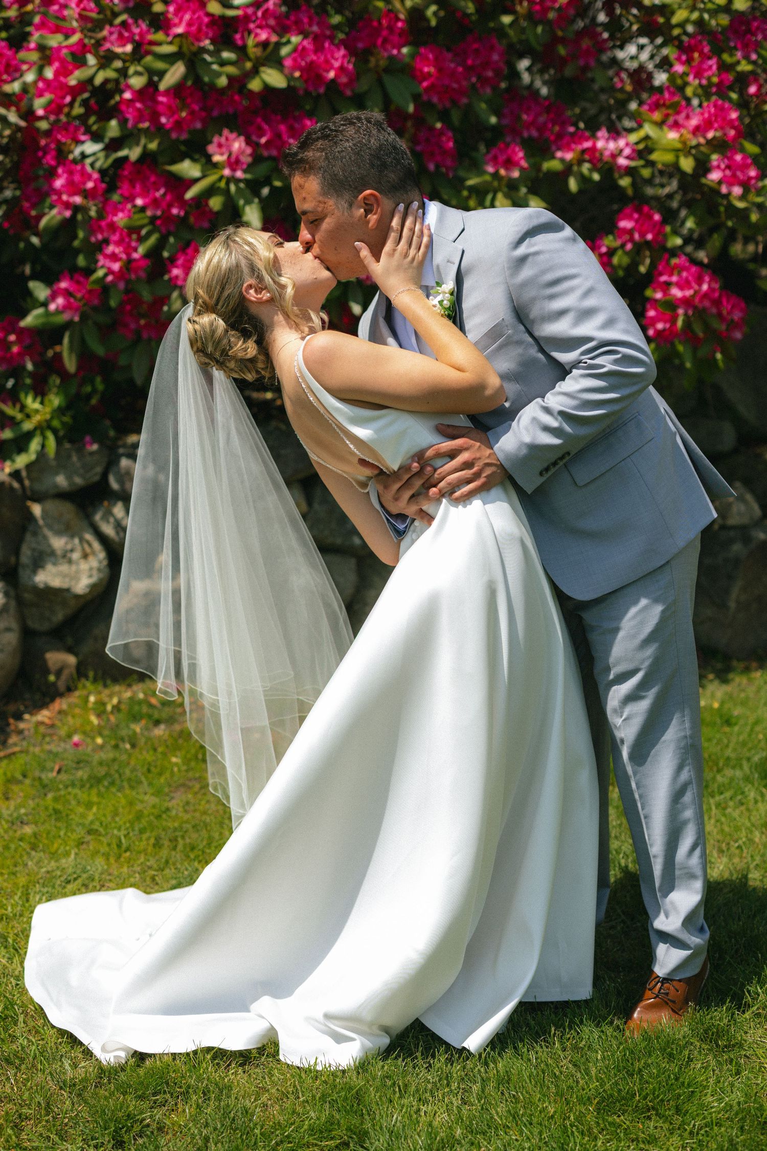 Bride and groom sharing a romantic kiss outdoors, surrounded by vibrant pink flowers and lush greenery on their wedding day.