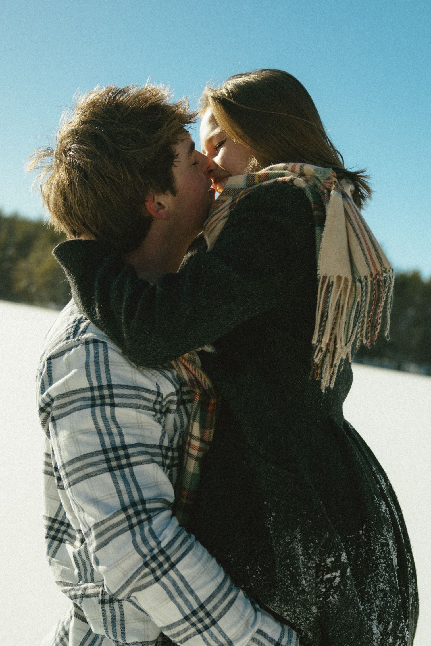 A couple shares a romantic winter kiss outdoors with snow in the background, wearing cozy winter clothing and scarves.