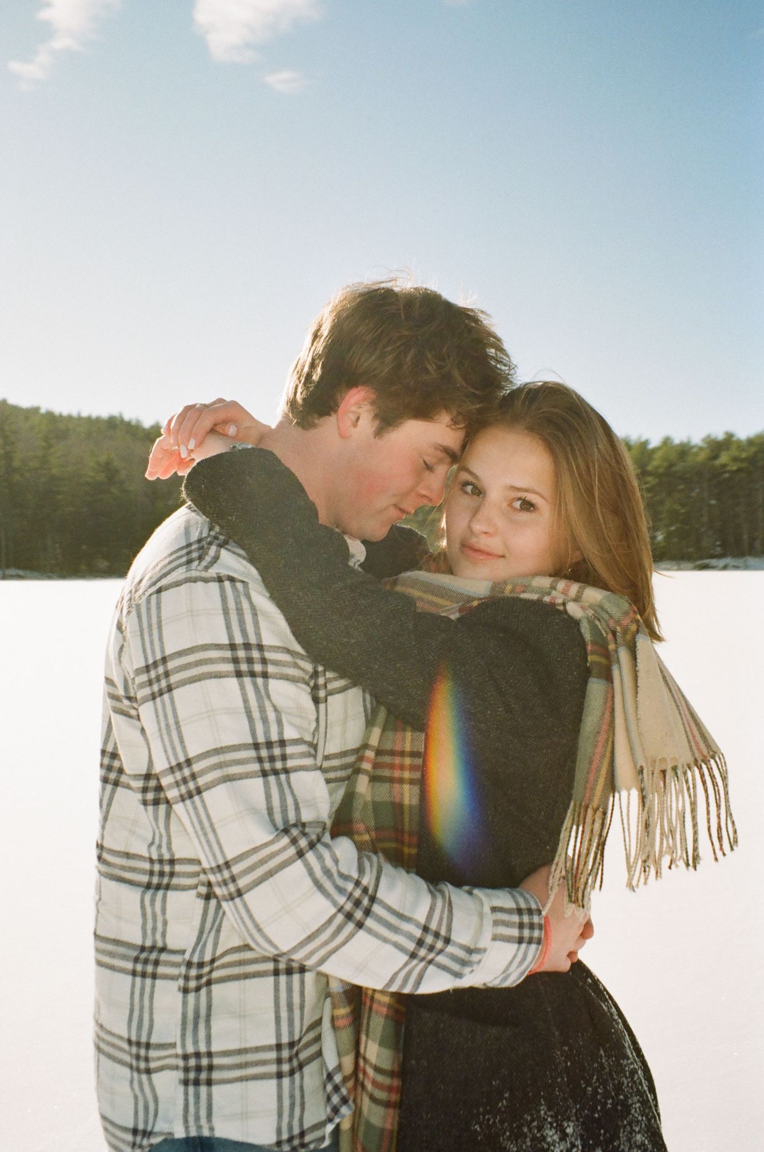 A romantic winter scene shows a couple embracing in the snow with a plaid jacket and scarf against a bright sky.